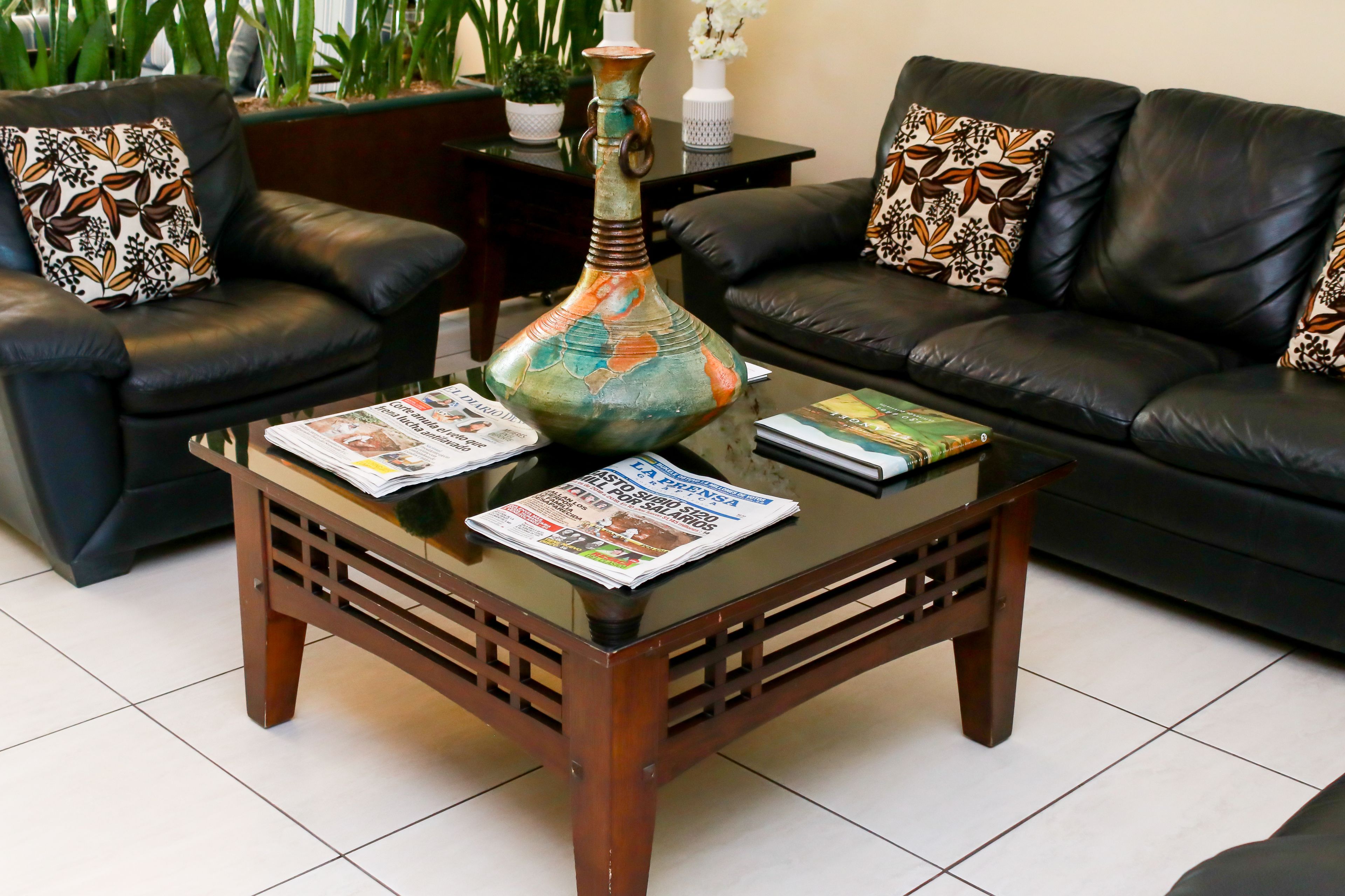 Un salon avec une table basse en verre, deux canapés en cuir noir ornés de coussins à motifs, un vase décoratif et un magazine posé sur la table.
