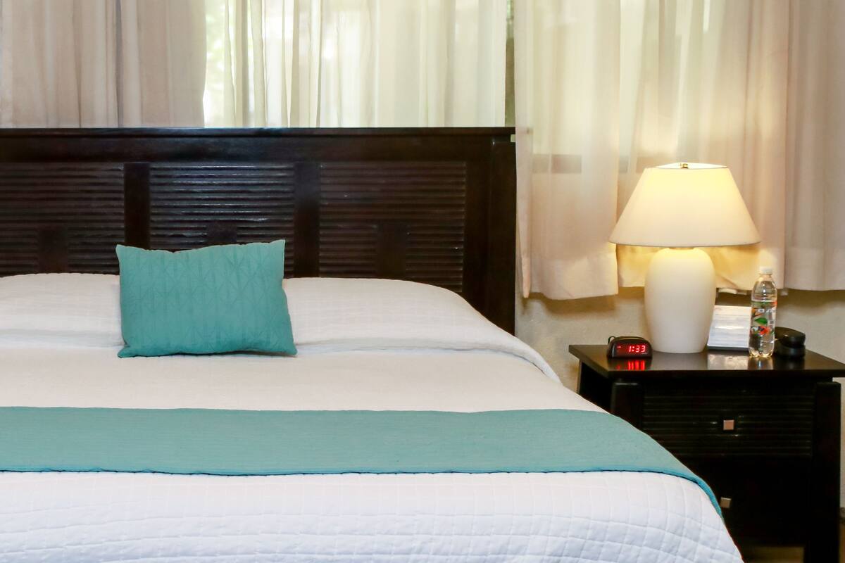 A neatly made bed with a teal pillow and a white comforter, a bedside lamp, and a digital clock on a nightstand.