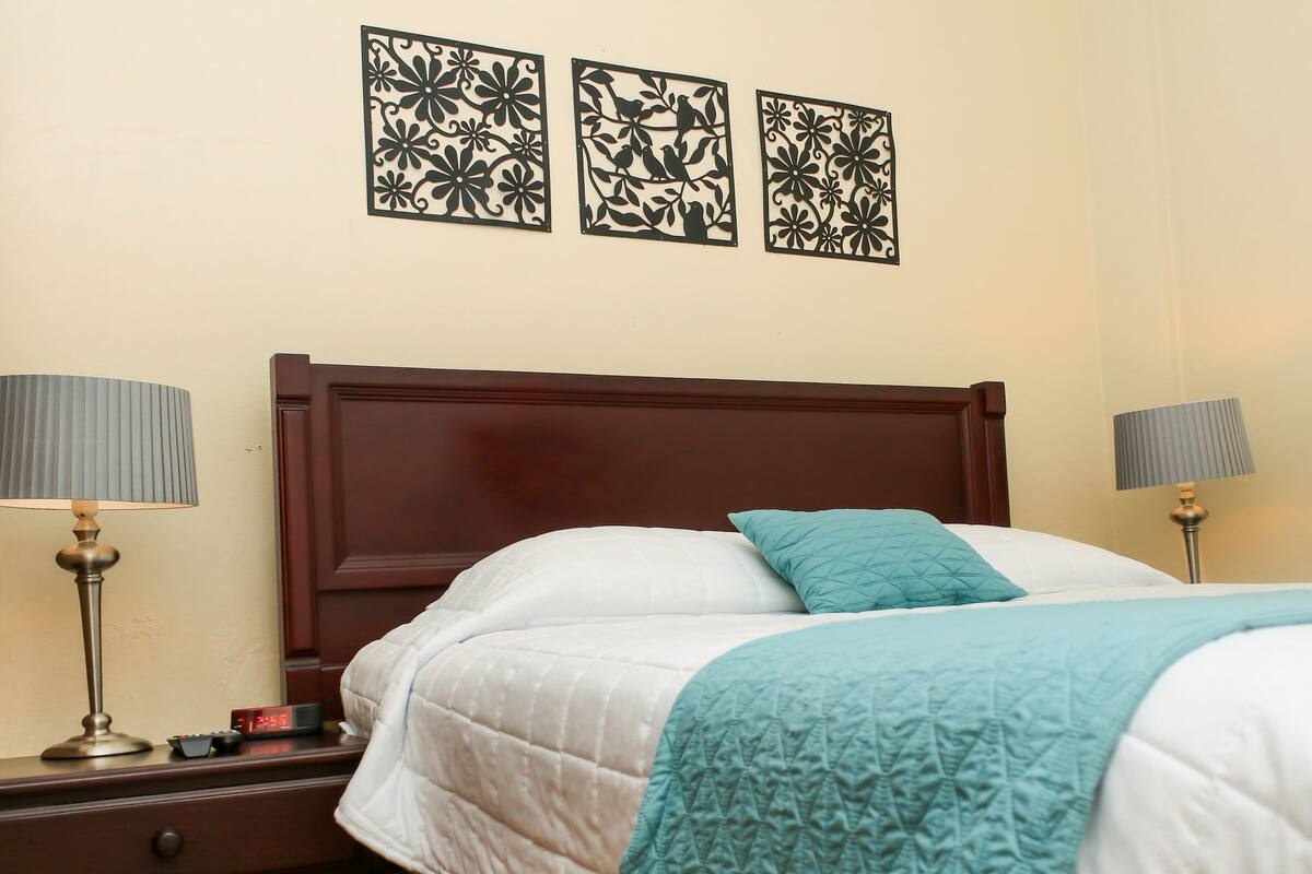 A neatly made bed with a white and teal blanket, flanked by two bedside tables with lamps, and three wall-mounted decorative panels above.
