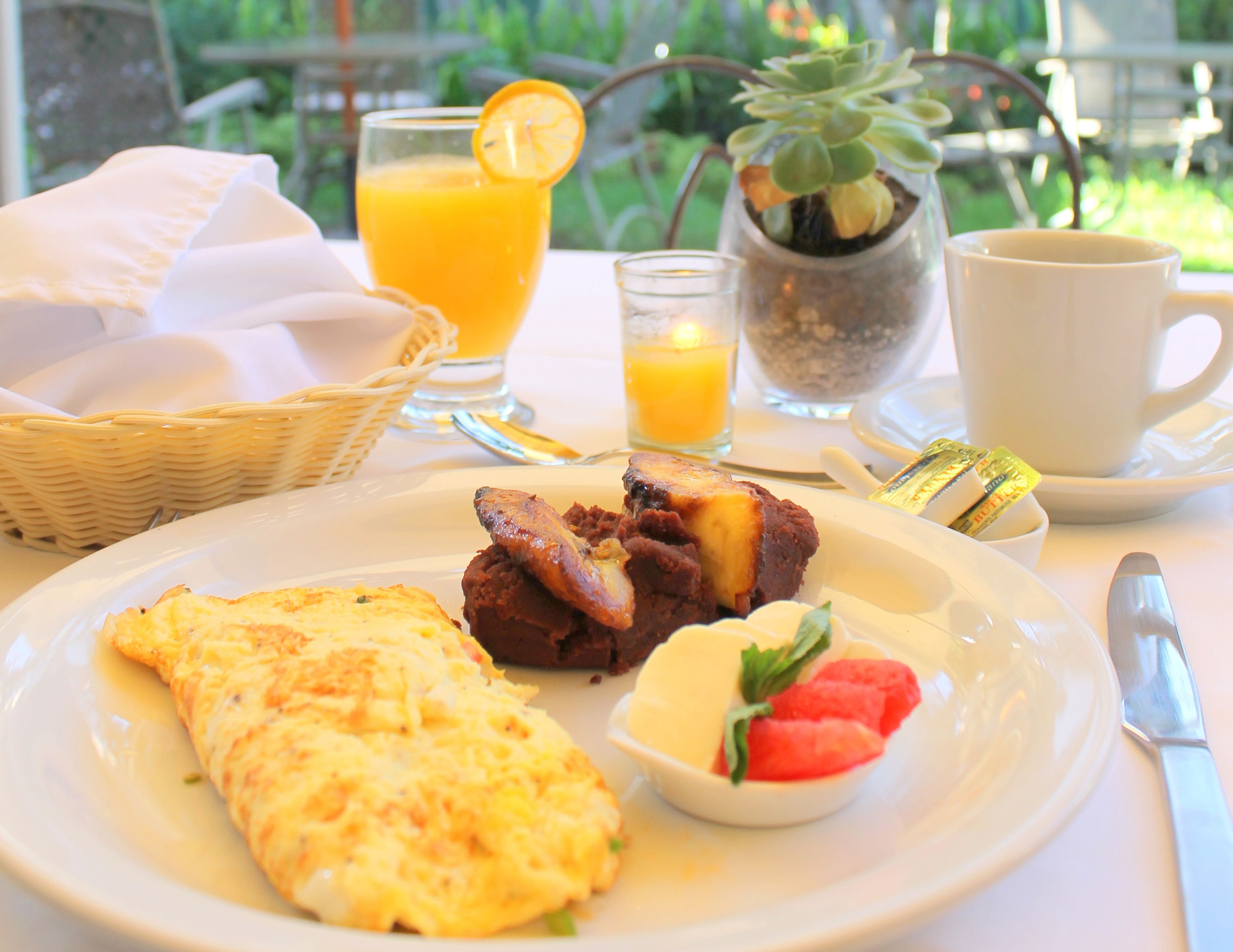 Une table mise pour le petit-déjeuner avec une assiette de nourriture, un verre de jus d’orange, une tasse et une petite plante en pot.