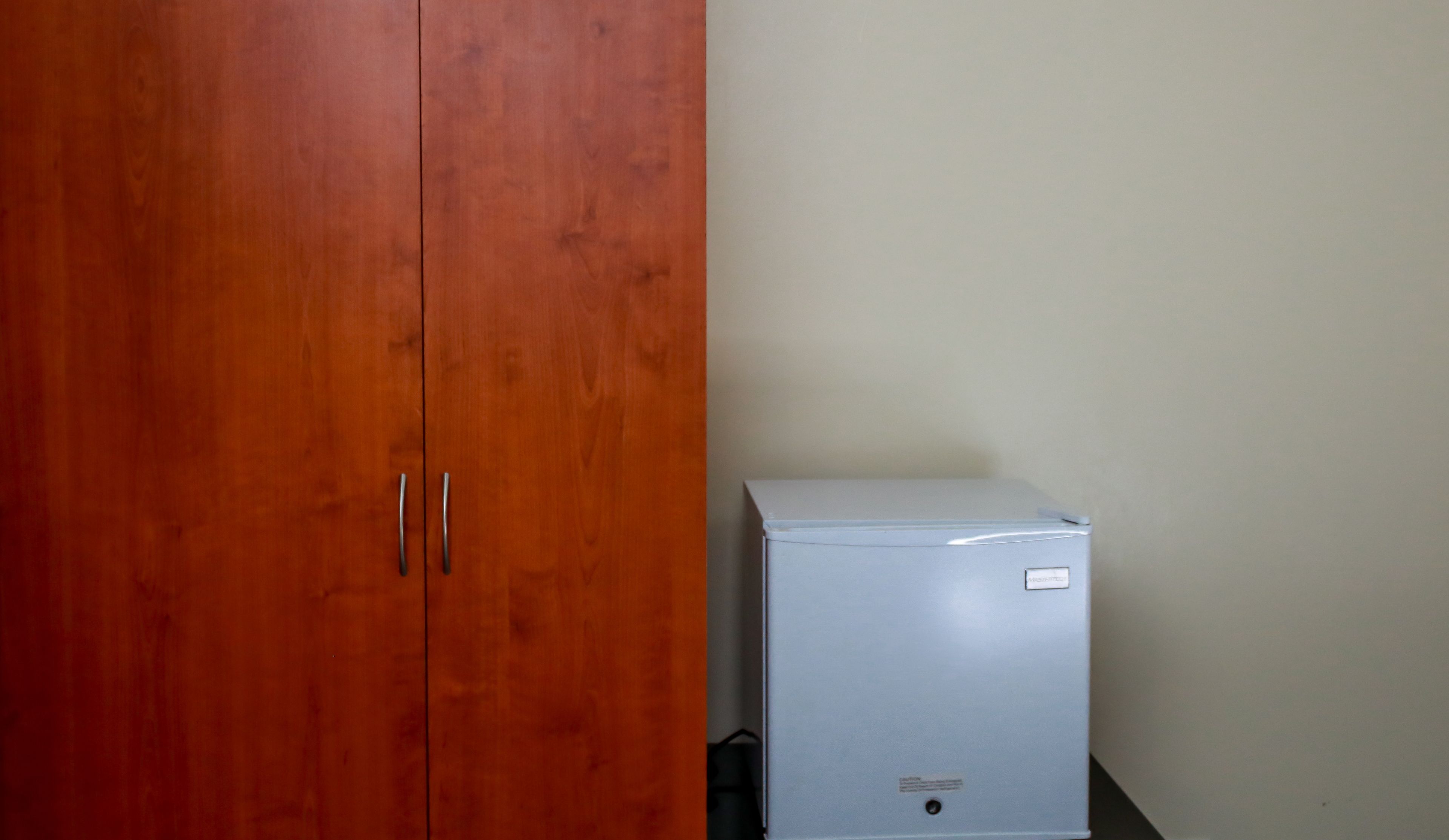 A room with a wooden wardrobe and a small white appliance, likely a refrigerator, against a plain wall.