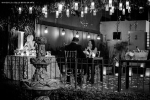 A black and white outdoor setting with a fountain, a table with chairs, and hanging lights.