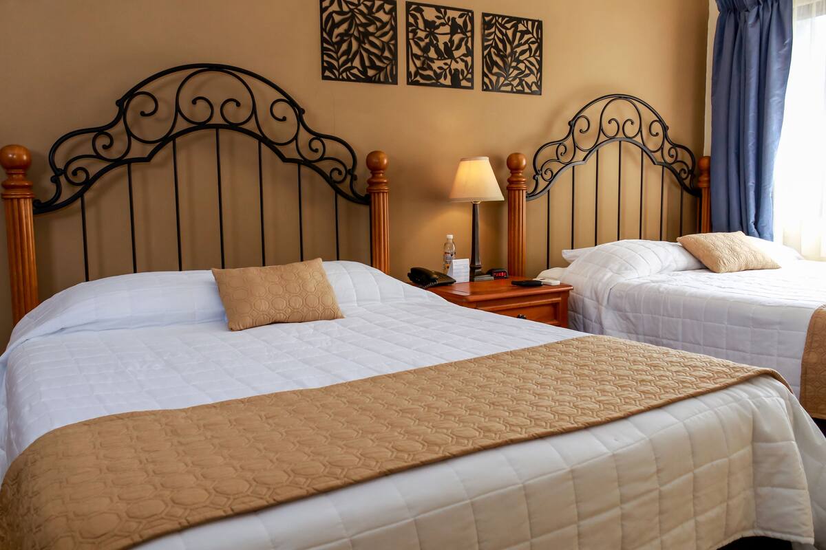 A hotel room with two beds, each with a wooden headboard and a beige quilted bedspread. There is a nightstand with a lamp and a phone between the beds.