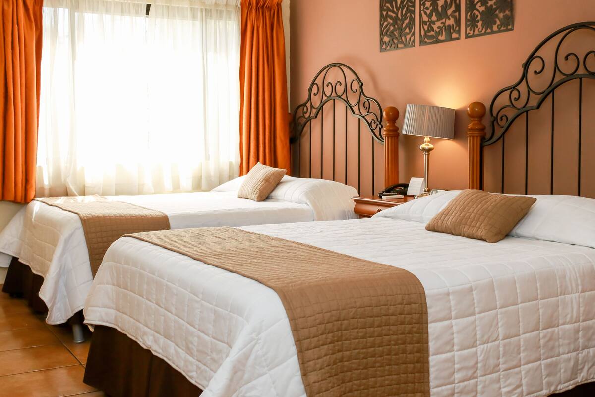 A hotel room with two beds, each with a white quilted bedspread and a brown throw blanket. There is a nightstand with a lamp and a telephone between the beds.