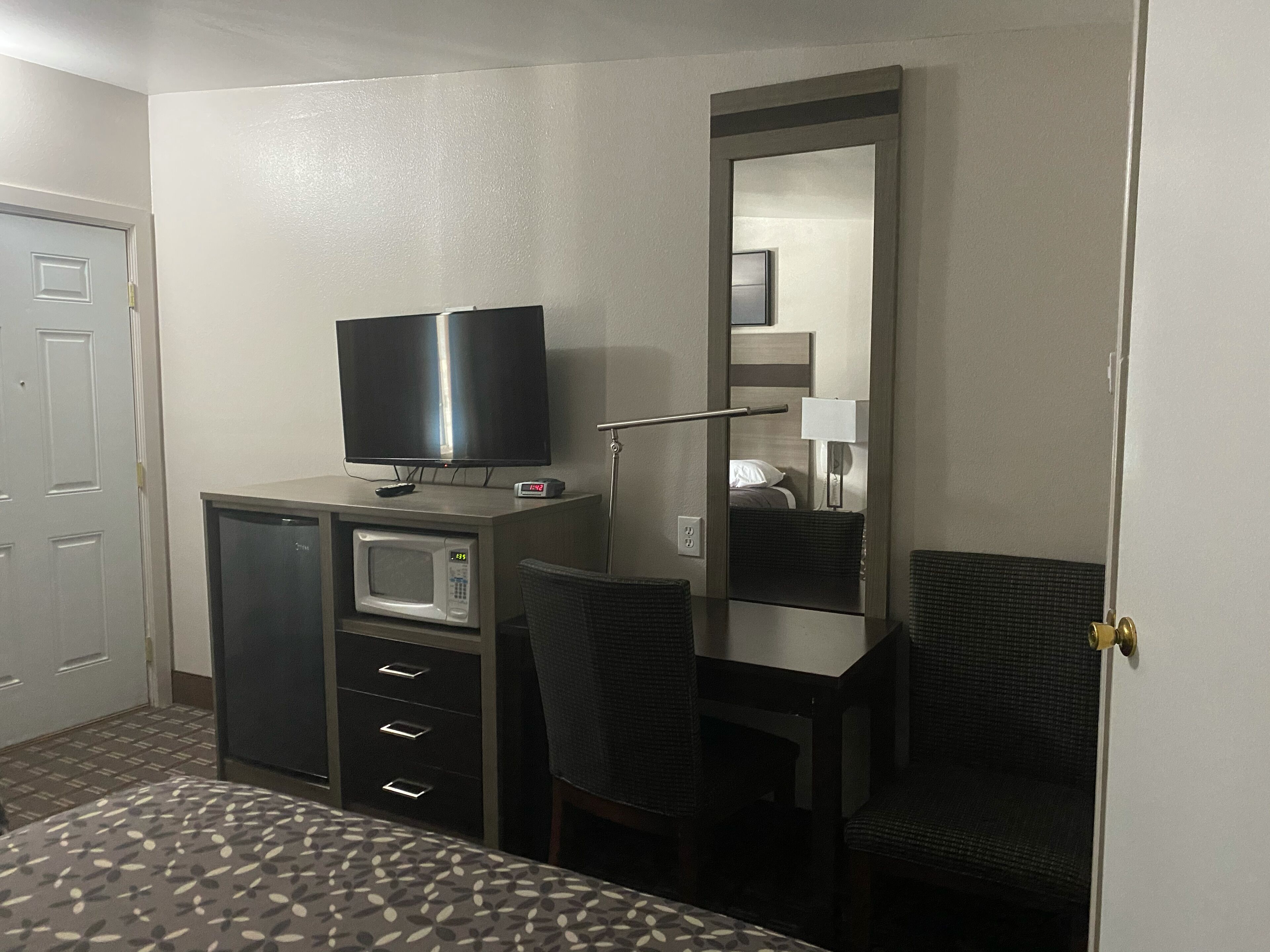 Una habitación de hotel con cama, escritorio, silla, televisor, microondas y espejo.