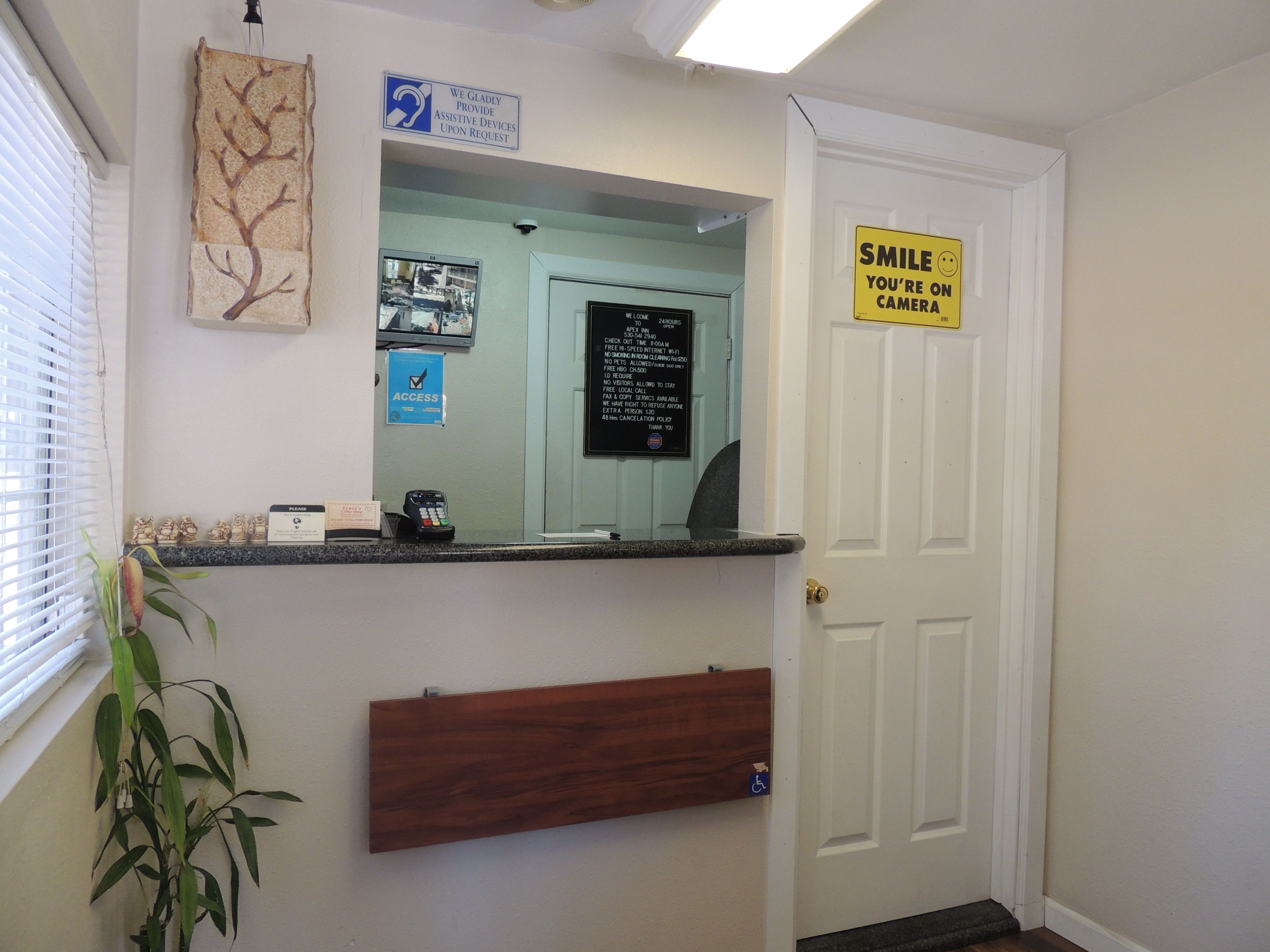 A reception area with a counter, a mirror, a sign offering assistive devices, a 'Smile, you're on camera' sign, a security camera, and a plant.