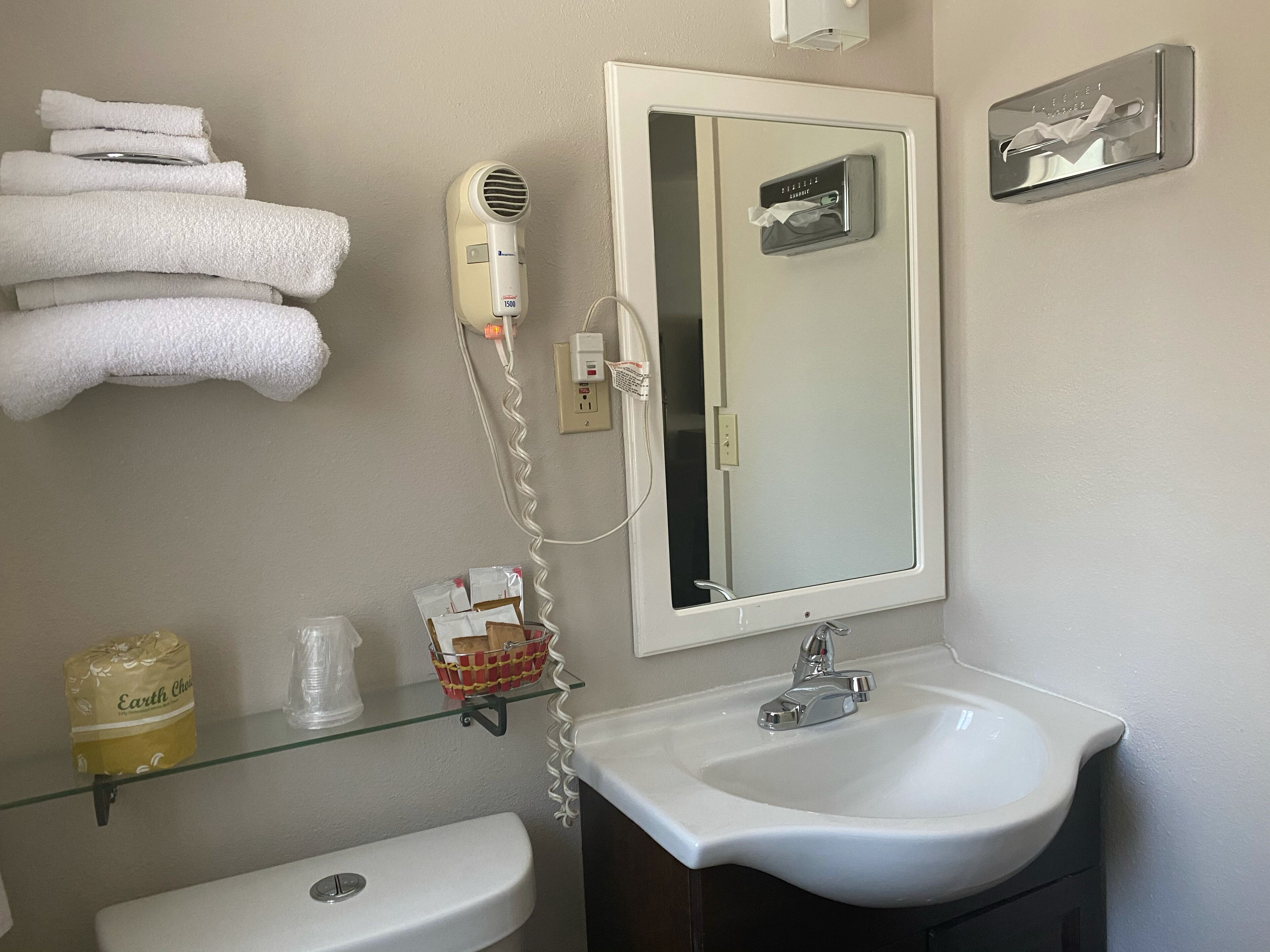 Un bagno con un lavandino bianco, uno specchio, un portasciugamani con asciugamani piegati, un asciugacapelli e uno scaffale in vetro con articoli da toilette.