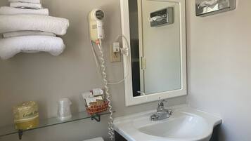 Un bagno con un lavandino bianco, uno specchio, un portasciugamani con asciugamani piegati, un asciugacapelli e uno scaffale in vetro con articoli da toilette.