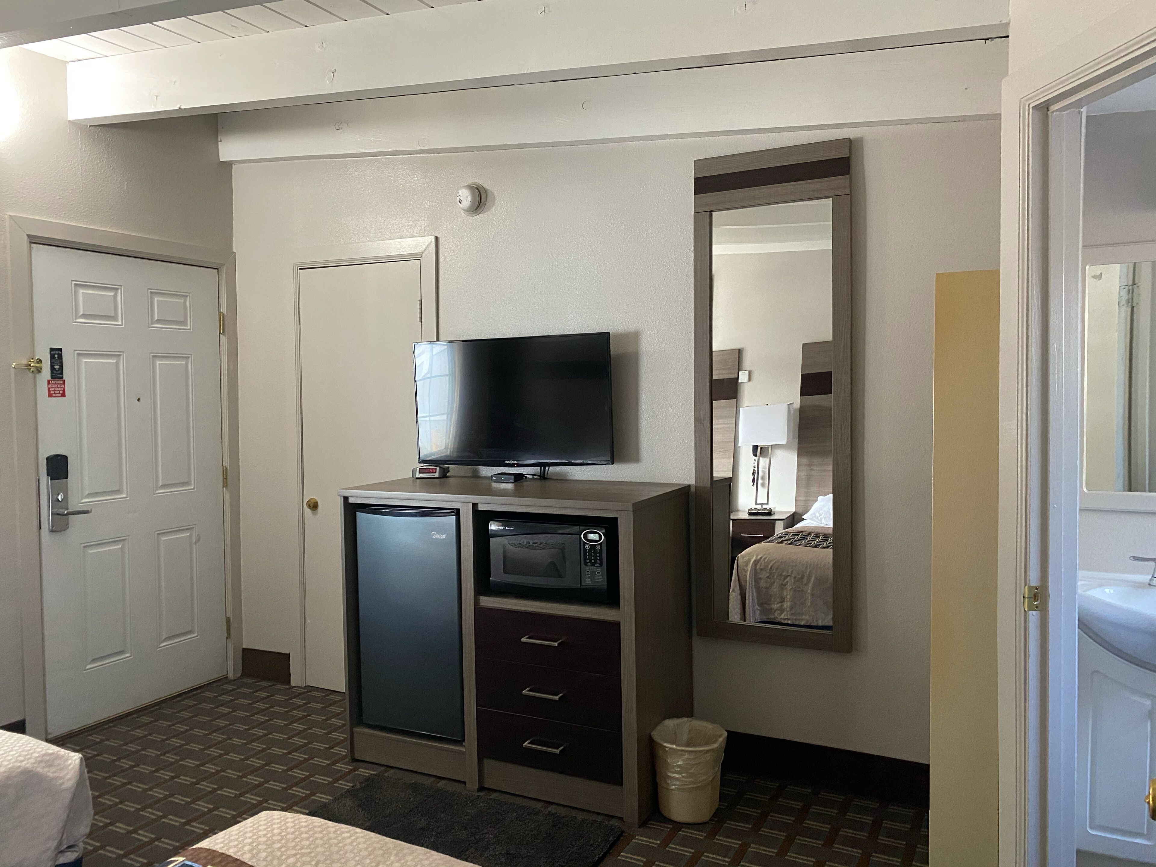 A hotel room with a television on a stand, a mini-fridge, a trash can, and a mirror on the wall.