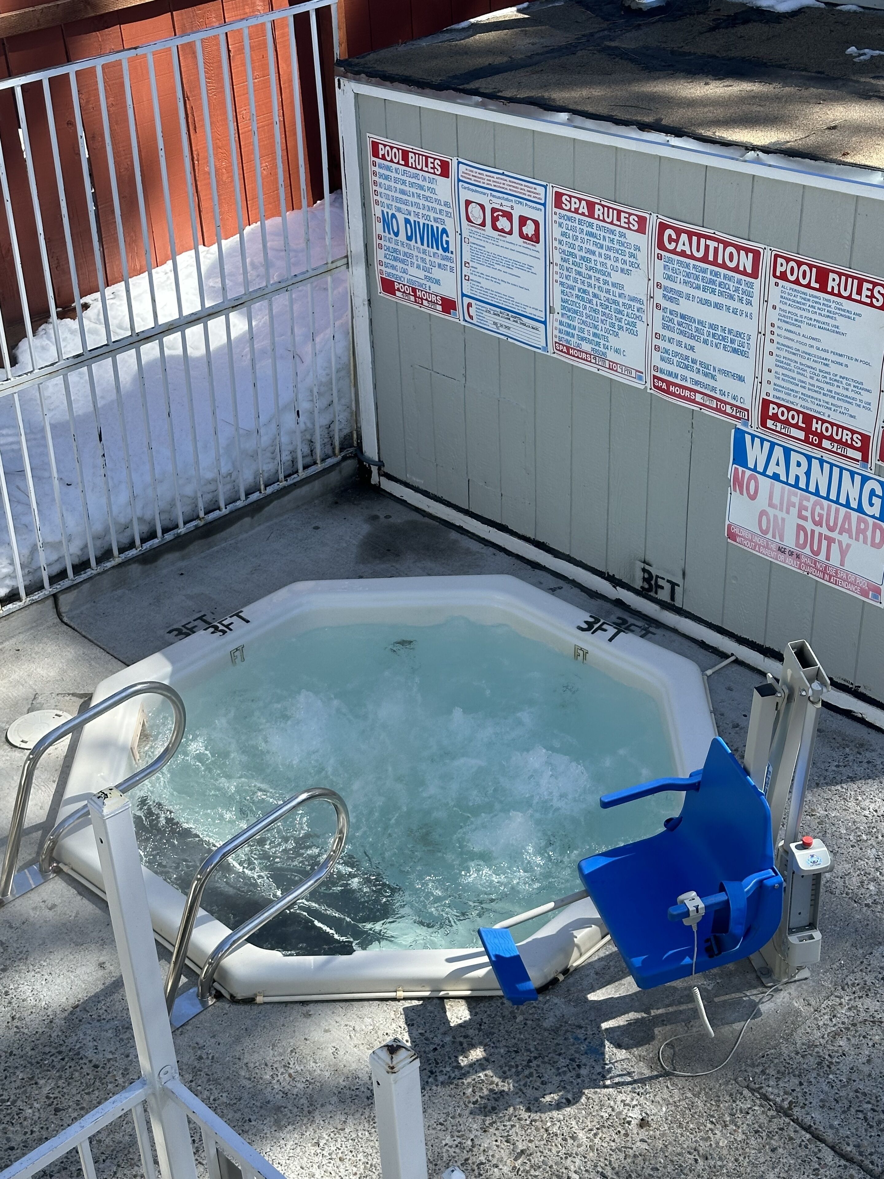 Un jacuzzi con baranda metálica y una silla azul, rodeado de carteles con normas de la piscina y el spa.