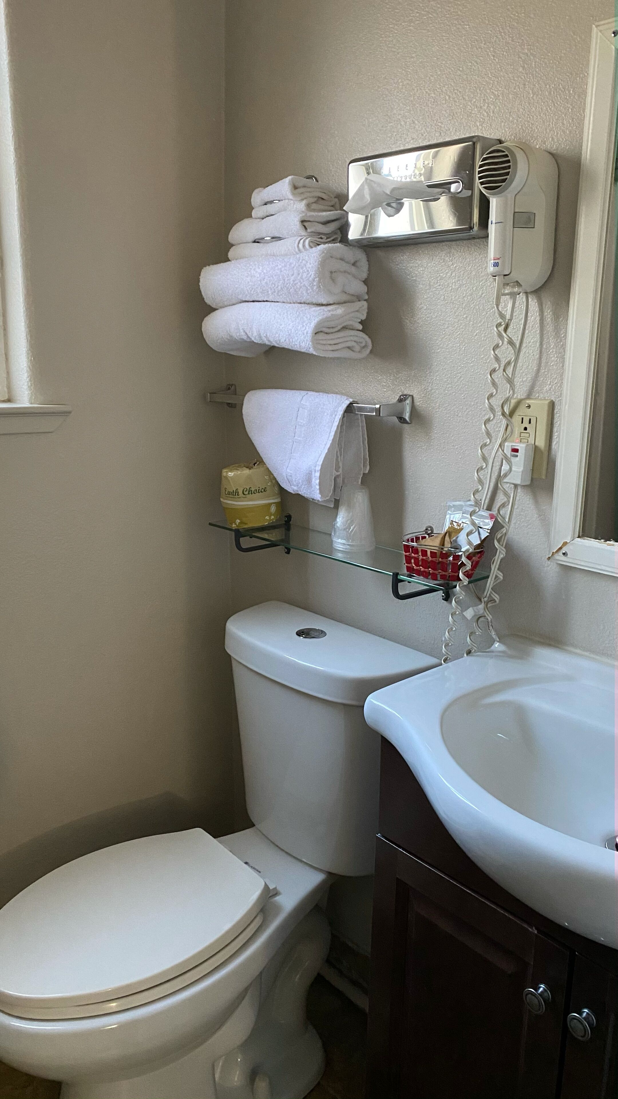 A bathroom with a toilet, a sink, a towel rack with folded towels, a hairdryer, and a glass shelf with toiletries.