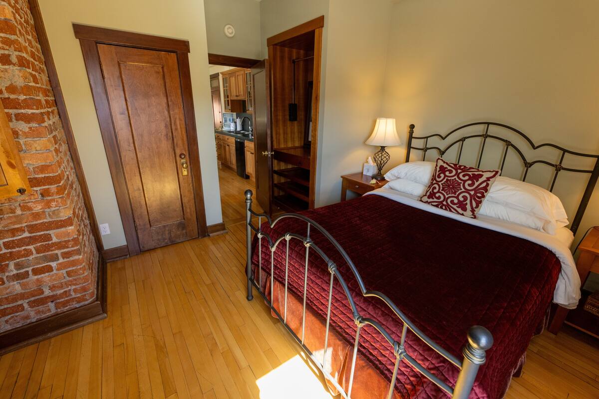 A bedroom with a wooden floor, a metal bed frame, a red bedspread, a bedside table with a lamp, and a door leading to a kitchen area.