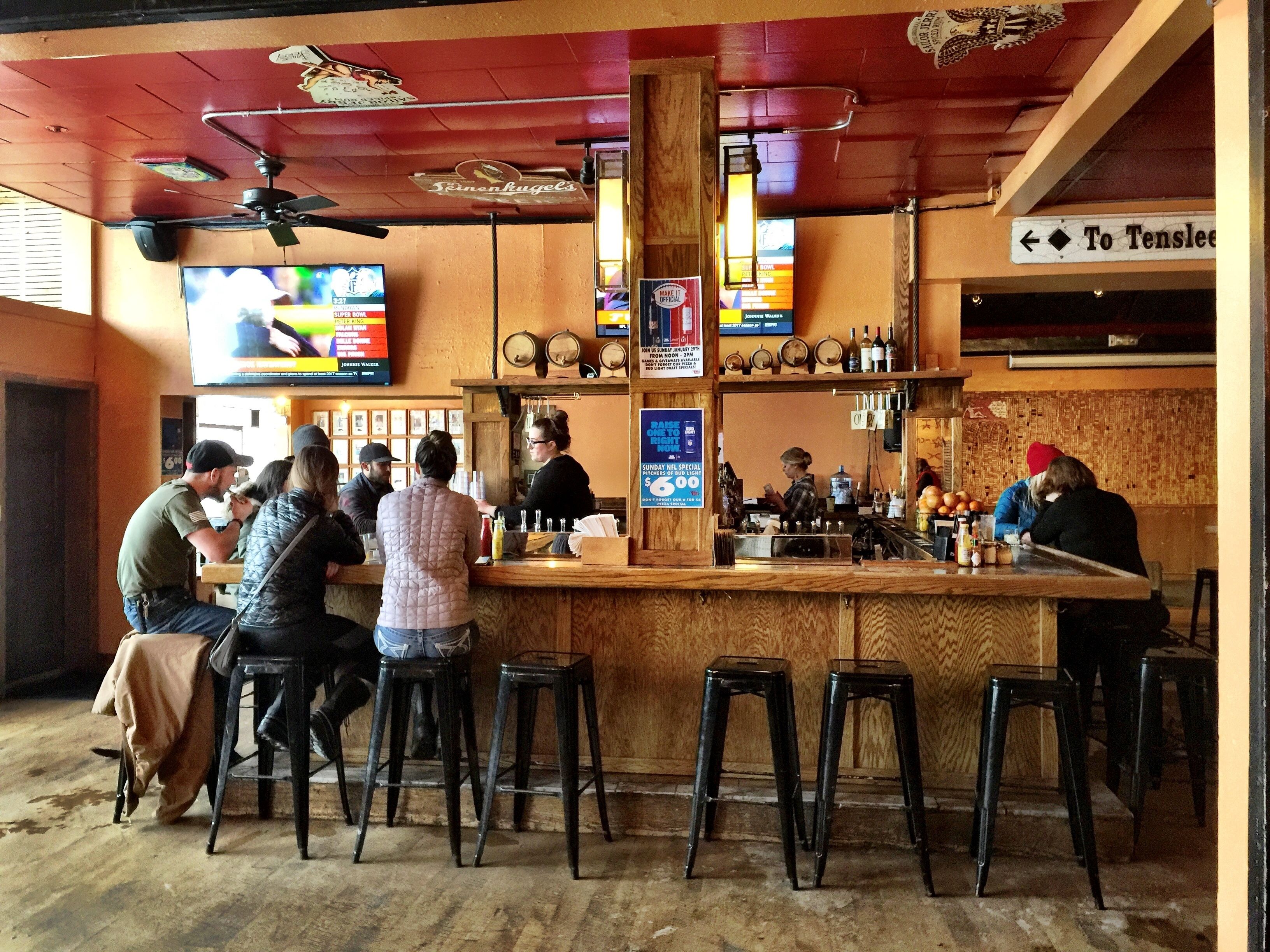 Un bar avec des tabourets de bar en bois, un comptoir où sont exposées des bouteilles et un téléviseur diffusant un événement sportif.