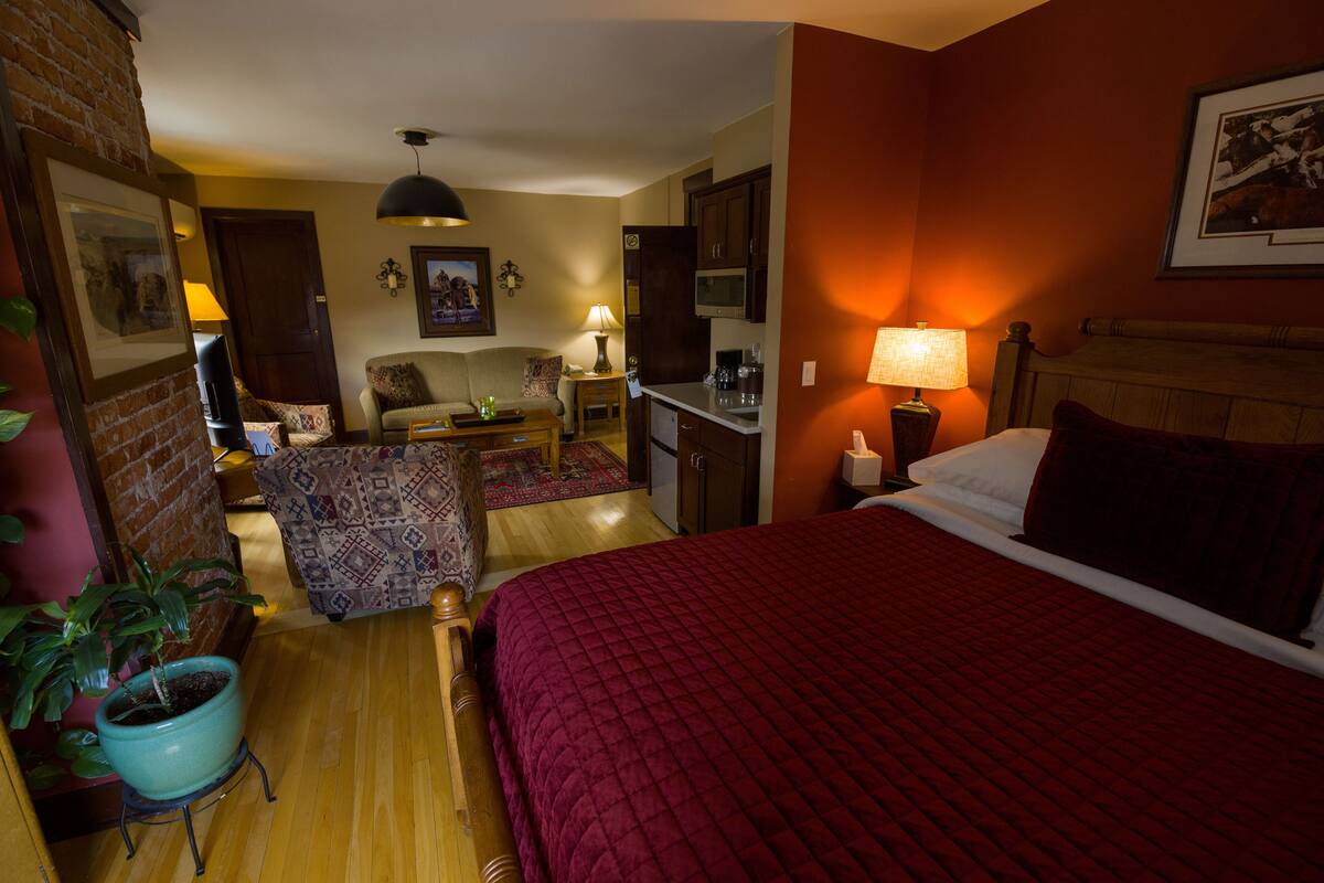 A hotel room with a large bed, a desk with a lamp, a chair, a small table, a potted plant, and a kitchen area.