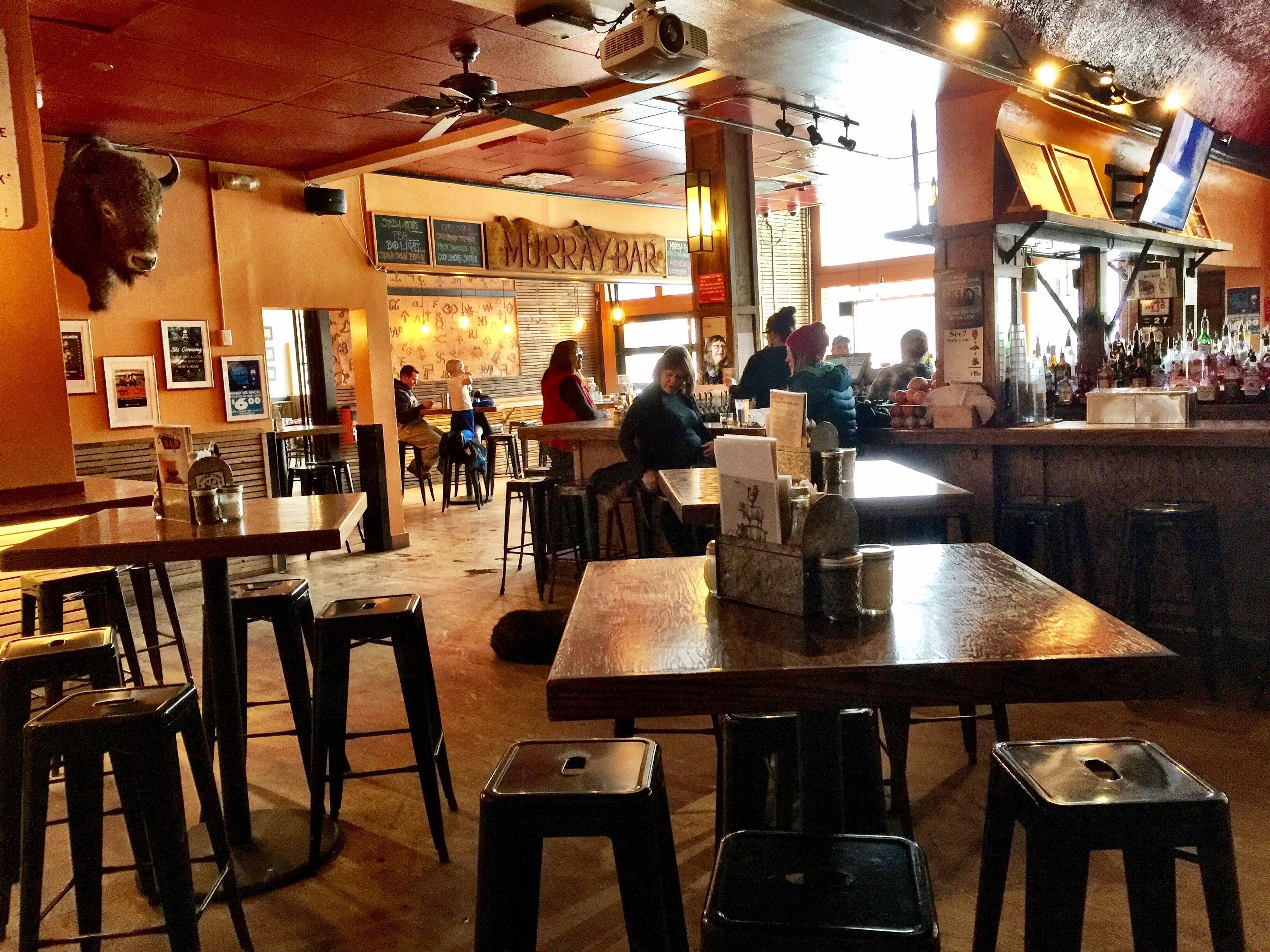 A restaurant interior with wooden tables and chairs, a bar area, and a ceiling fan.