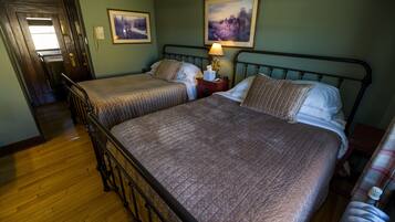 A room with two beds, wooden flooring, a nightstand with a lamp, and a door leading to a hallway.