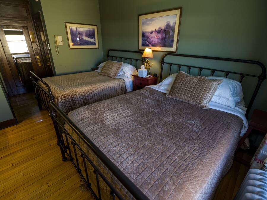 A room with two beds, wooden flooring, a nightstand with a lamp, and a door leading to a hallway.