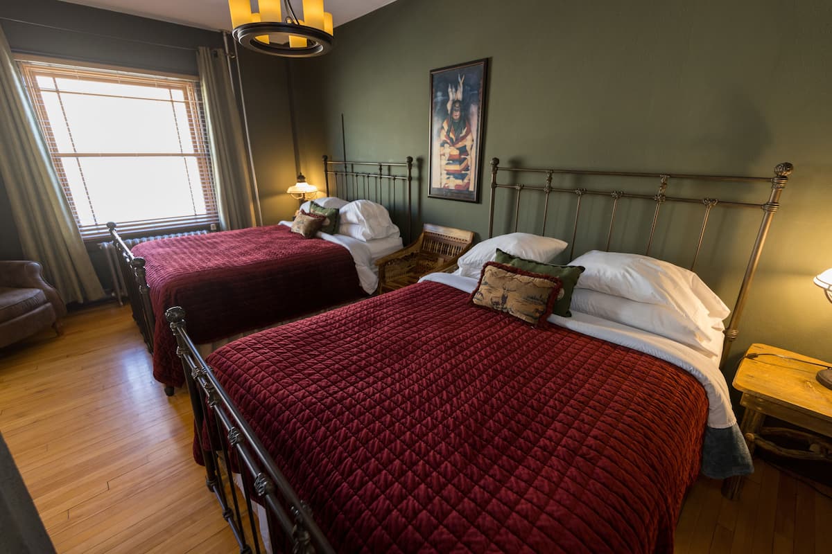 A room with two beds, wooden flooring, a window with curtains, a painting on the wall, and a nightstand with a lamp.