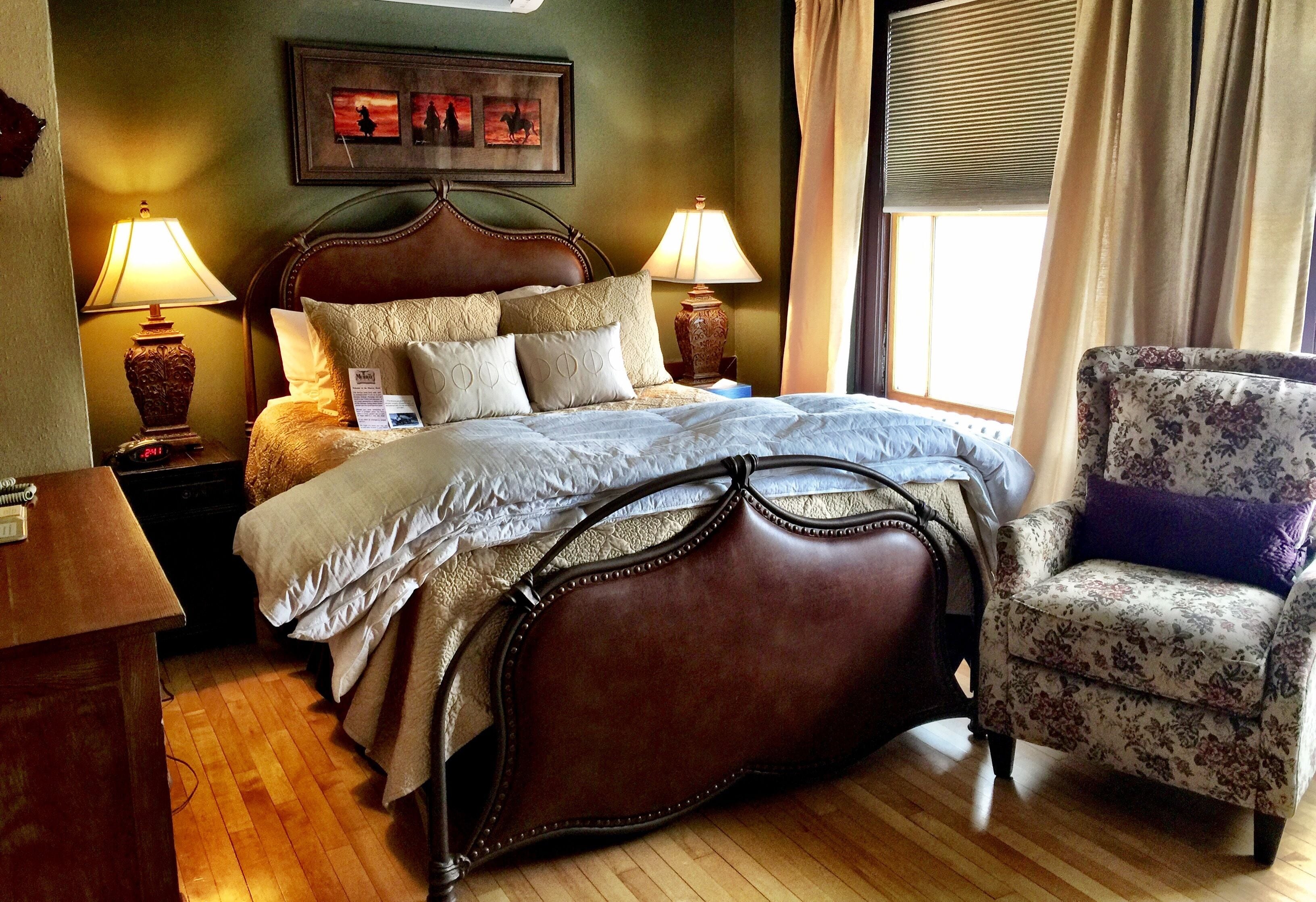 A neatly made bed with a headboard, two bedside lamps, a chair with a floral pattern, and a framed picture on the wall.