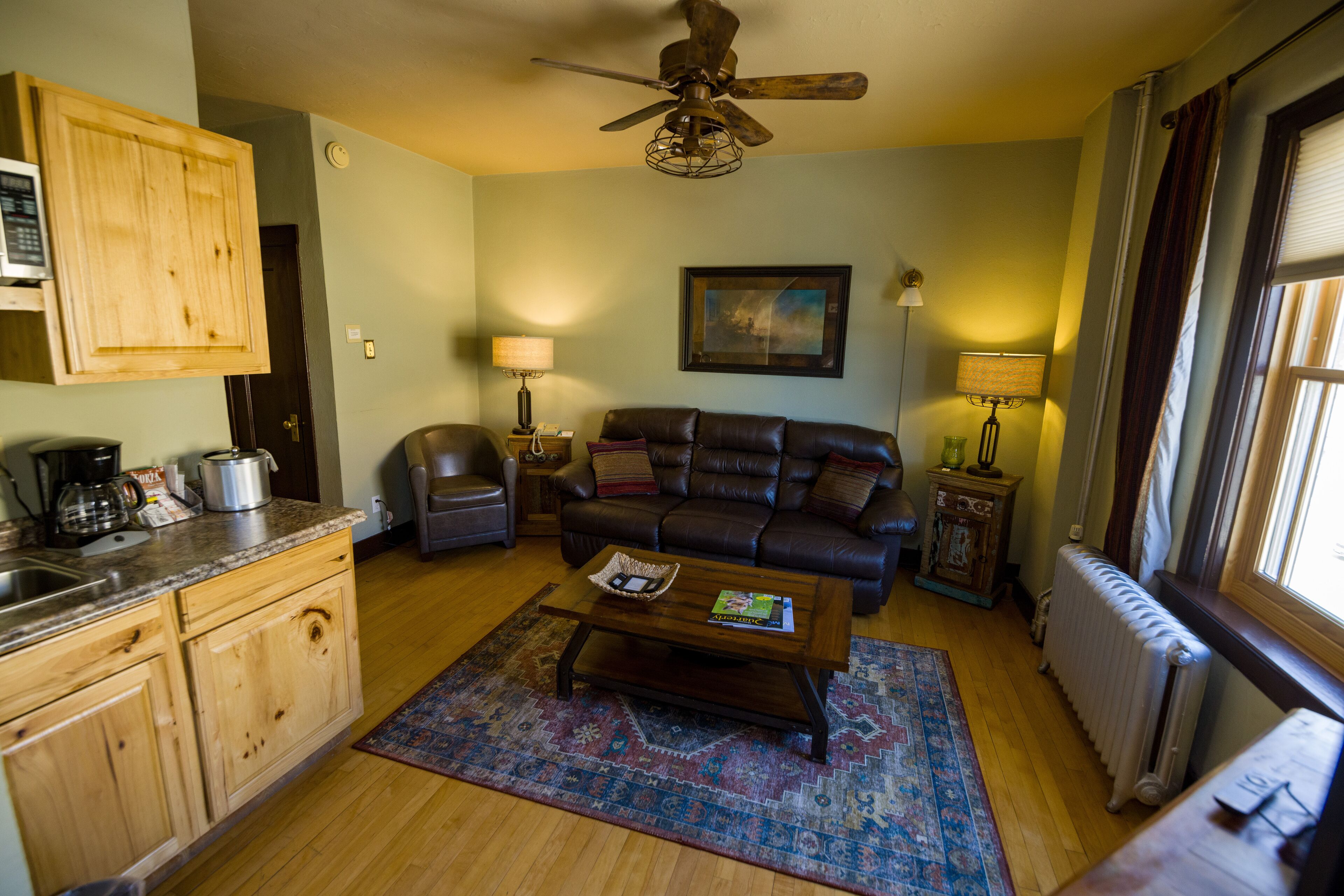 A living room with a sofa, coffee table, and a rug. There is a kitchen area with cabinets and a stove. A ceiling fan is visible, and a window provides natural light.