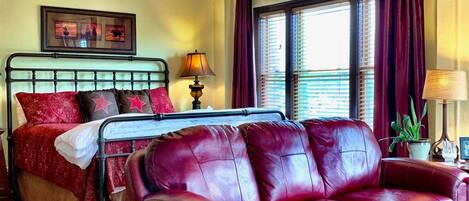A room with a bed, a red leather sofa, a wooden floor, a window with curtains, and a wall-mounted lamp.