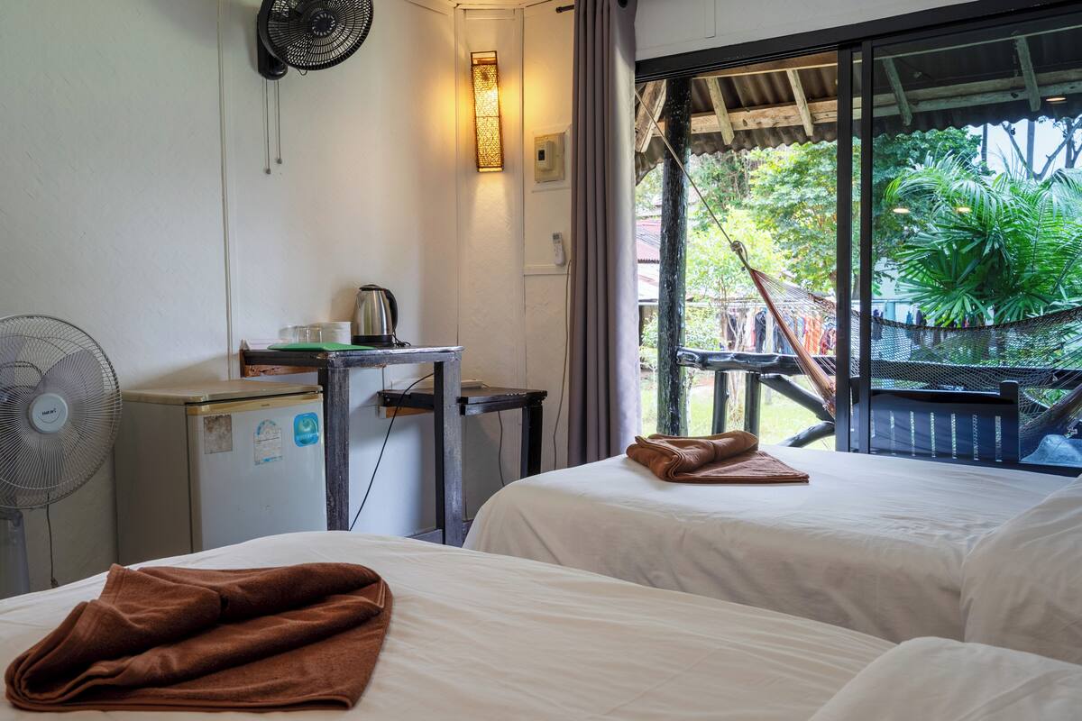 A hotel room with two beds, a desk with a kettle, a fan, and a view of the outdoors.