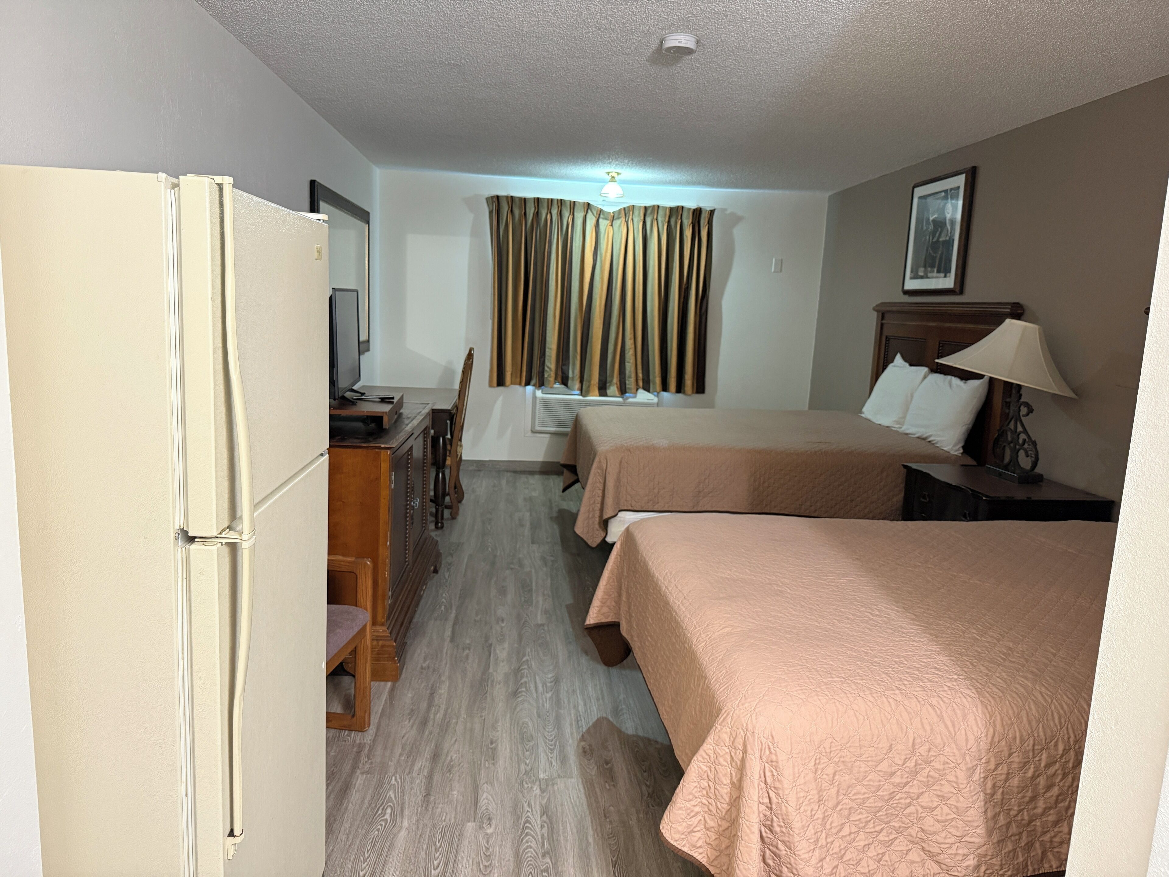 Een hotelkamer met twee bedden, een bureau met stoel, een koelkast, een televisie en een raam met gordijnen.