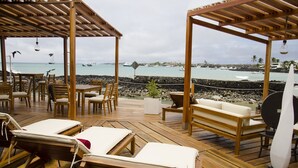 A wooden deck with lounge chairs and a table, overlooking a rocky shoreline with boats and a clear sky.