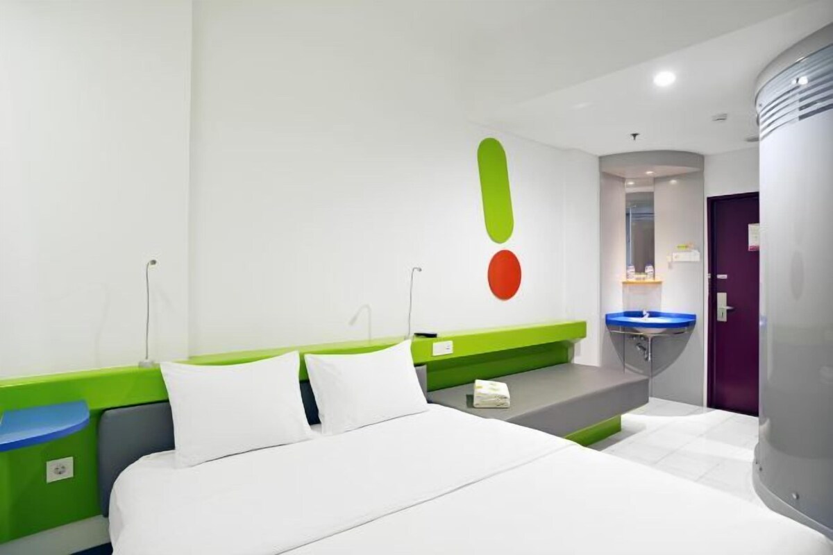Une chambre d’hôtel moderne avec un lit, un mur d’accent vert, une salle de bain avec un lavabo bleu et une porte violette.