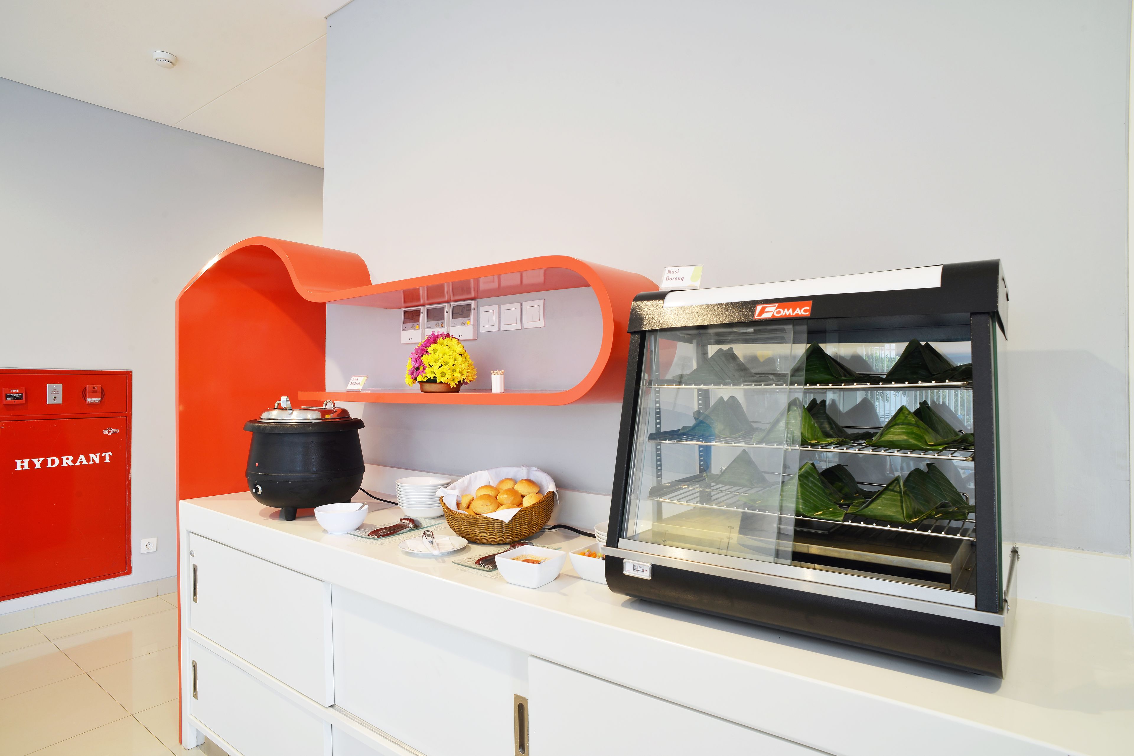 A breakfast buffet area with a food display case, a red cabinet labeled "HYDRANT," and a shelf with a yellow flower arrangement.