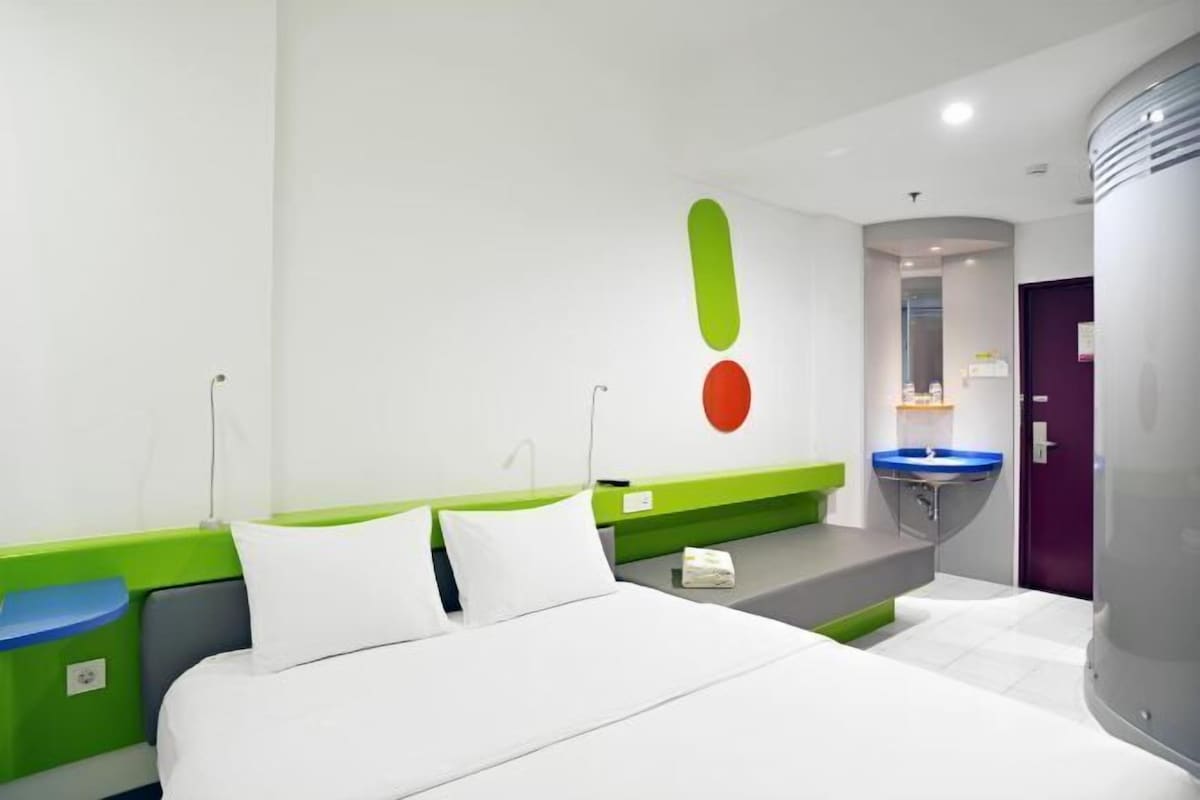 Une chambre d’hôtel moderne avec un lit, un mur d’accent vert, une salle de bain avec un lavabo bleu et une porte violette.