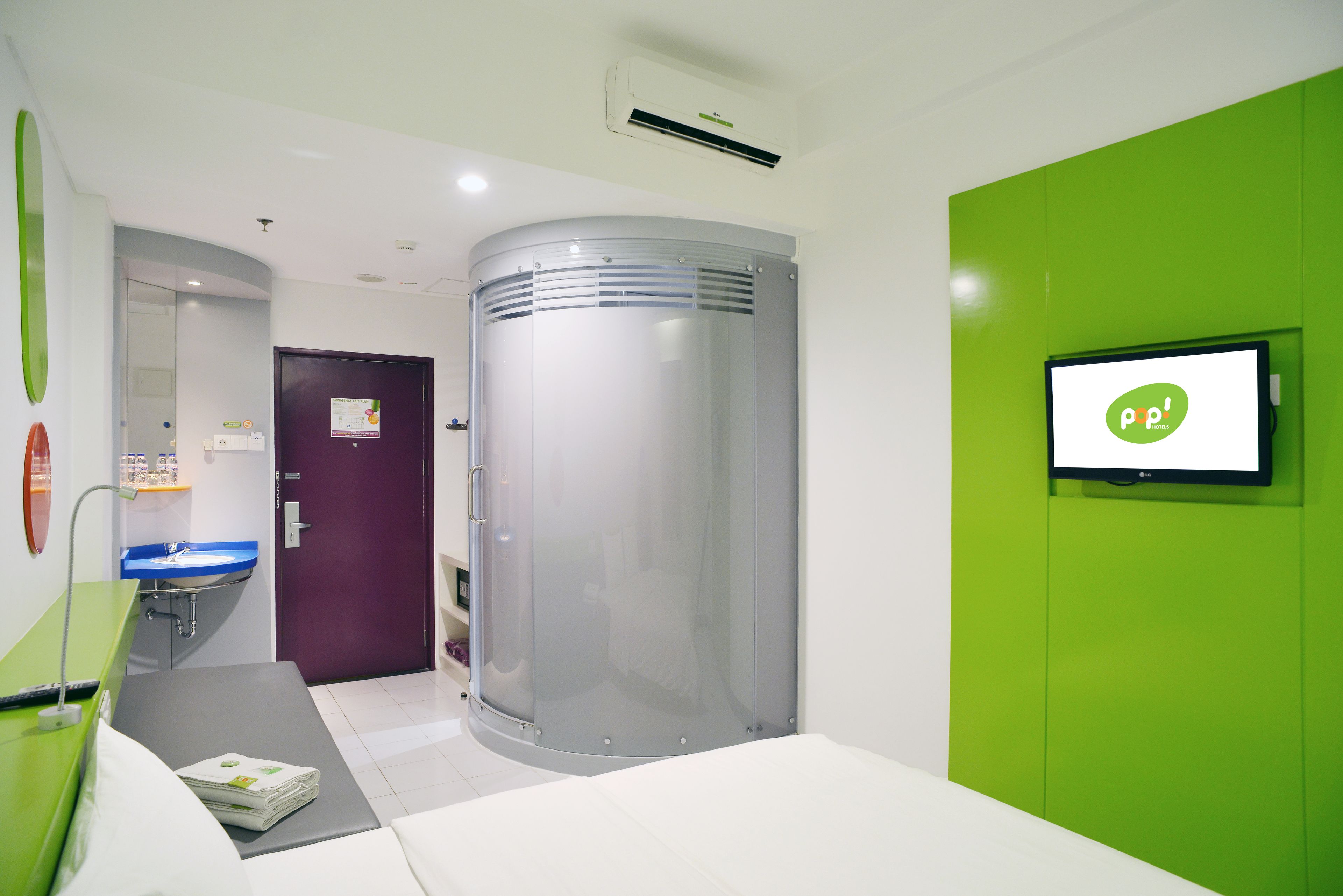 A modern hotel room with a curved glass partition, a green accent wall, a flat-screen TV, a purple door, and a white sink.
