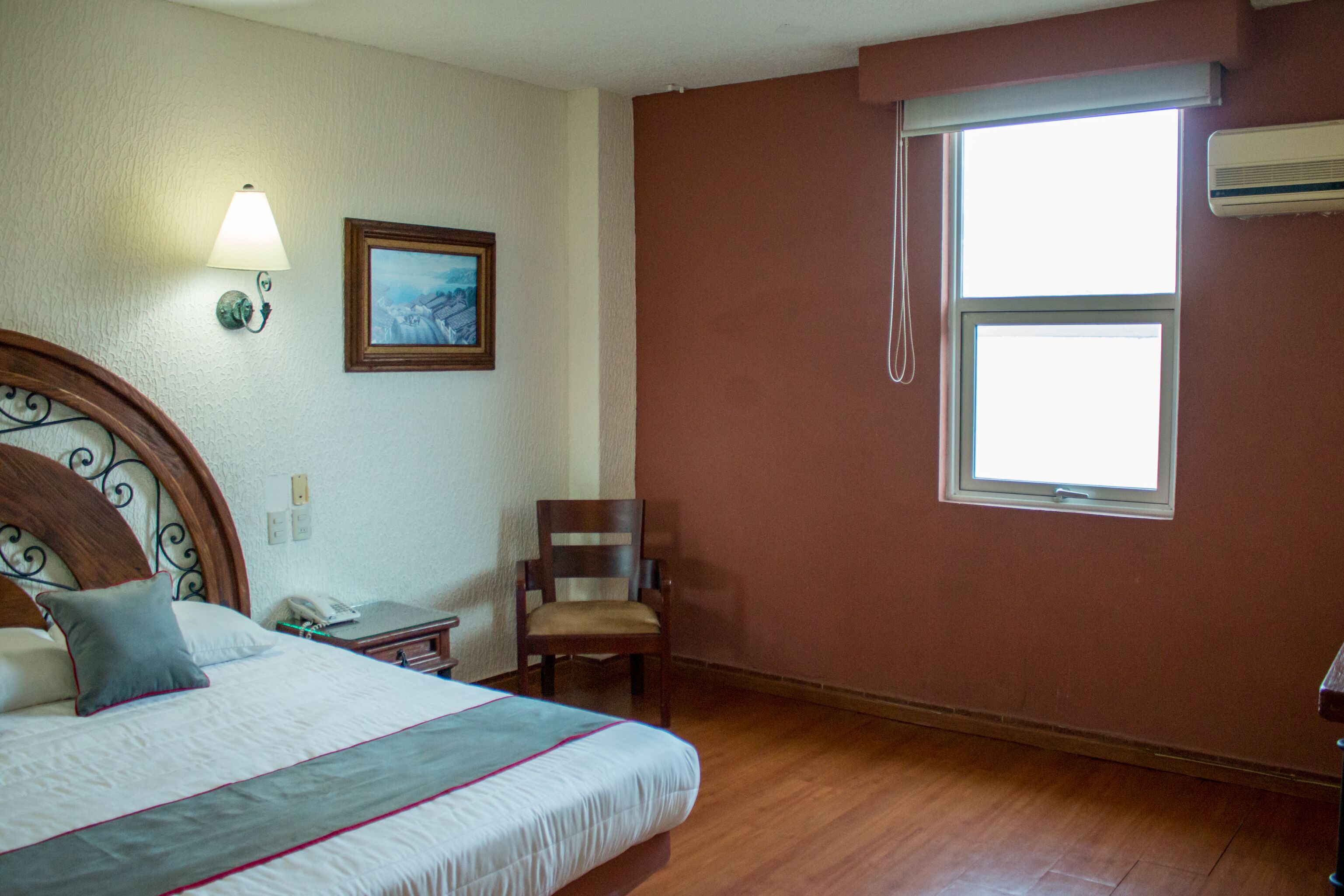 A hotel room with a bed, a chair, a desk, a lamp, a painting, and a window.