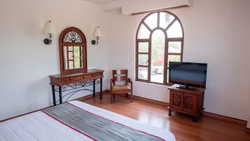 A bedroom with a bed, a wooden dresser, a television, and a window with a view of greenery.