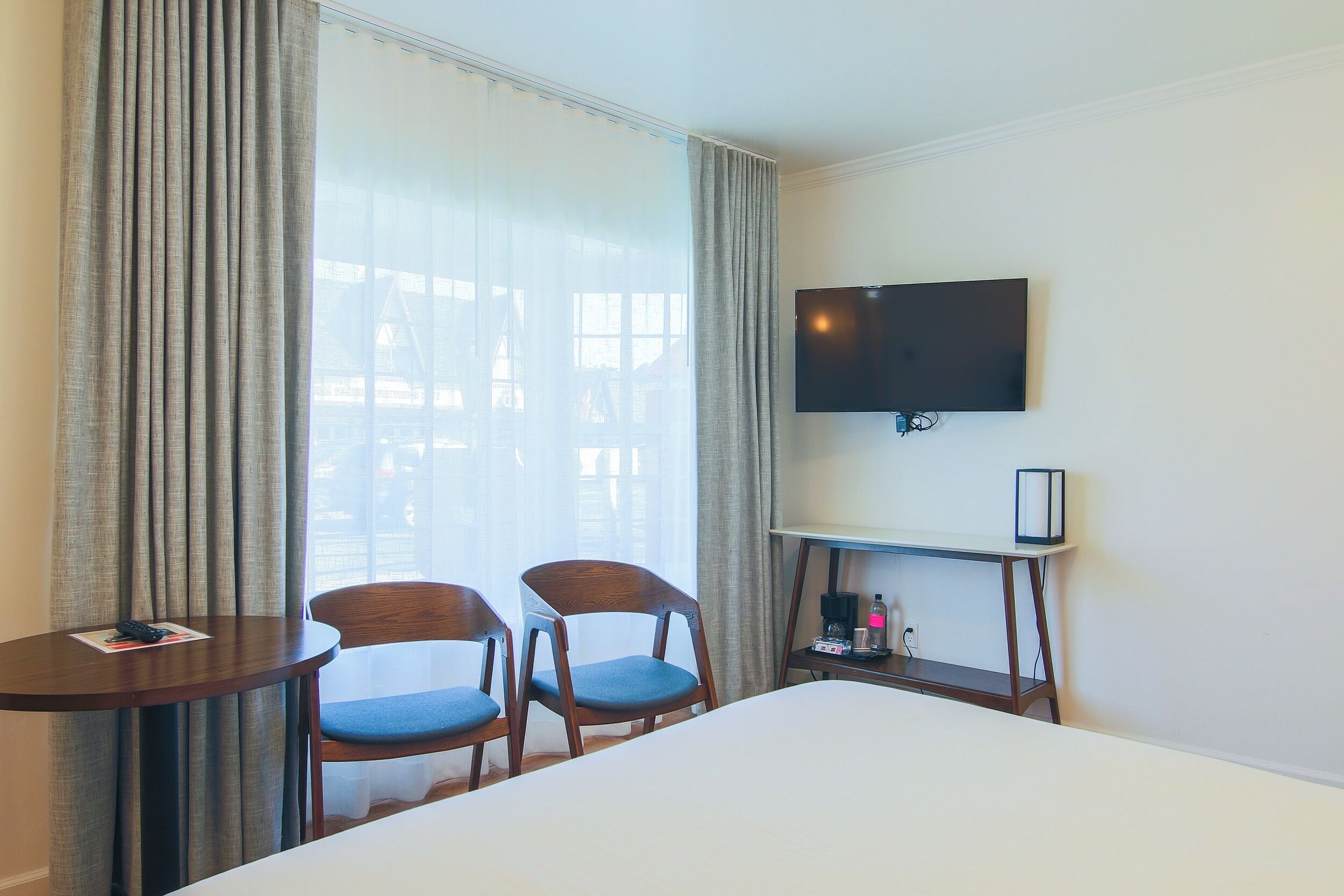 A hotel room with a bed, two chairs, a small table with a remote control, a TV on the wall, and a coffee maker on a side table.