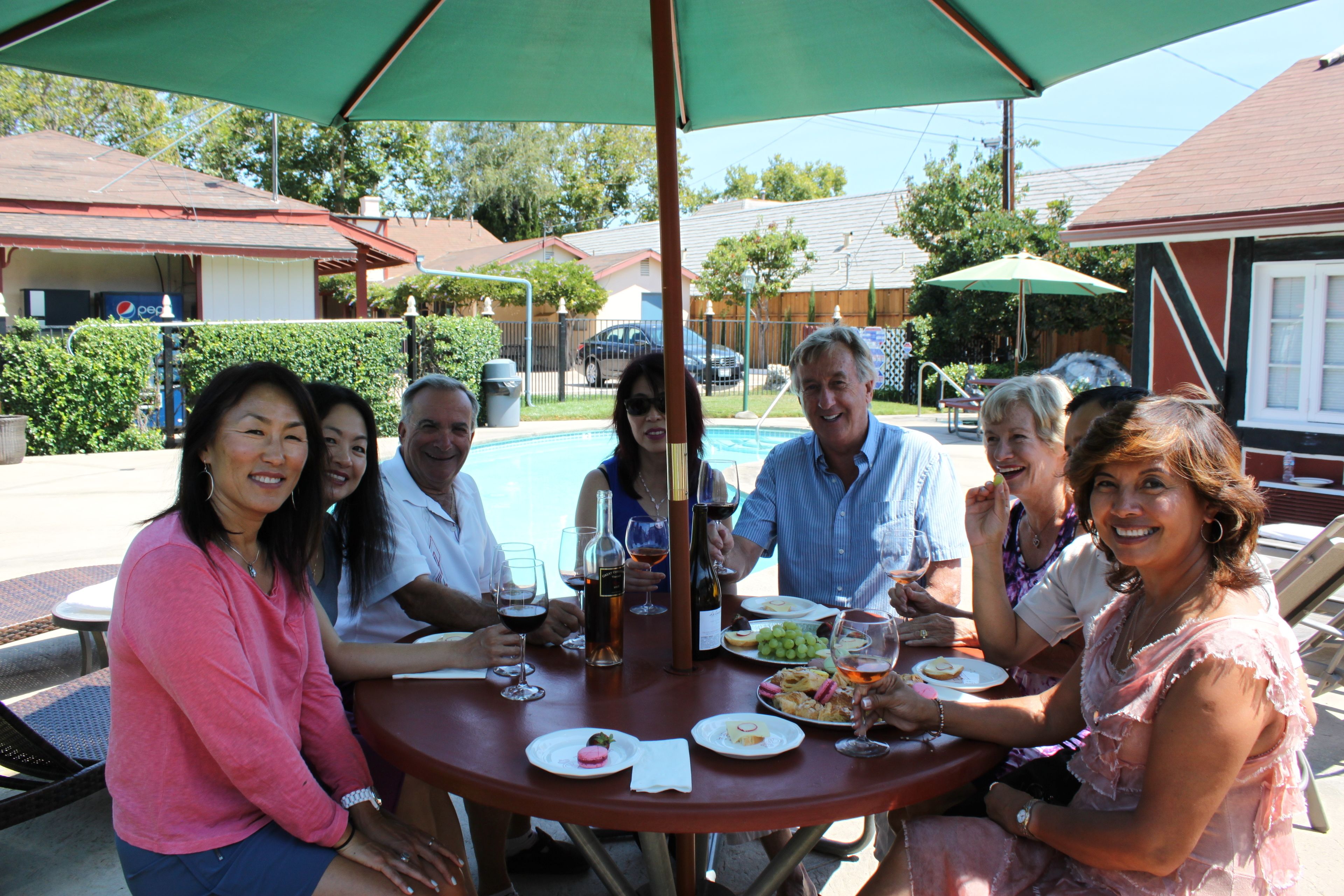 A group of people seated around a round table with a green umbrella, dining outdoors with a pool in the background.