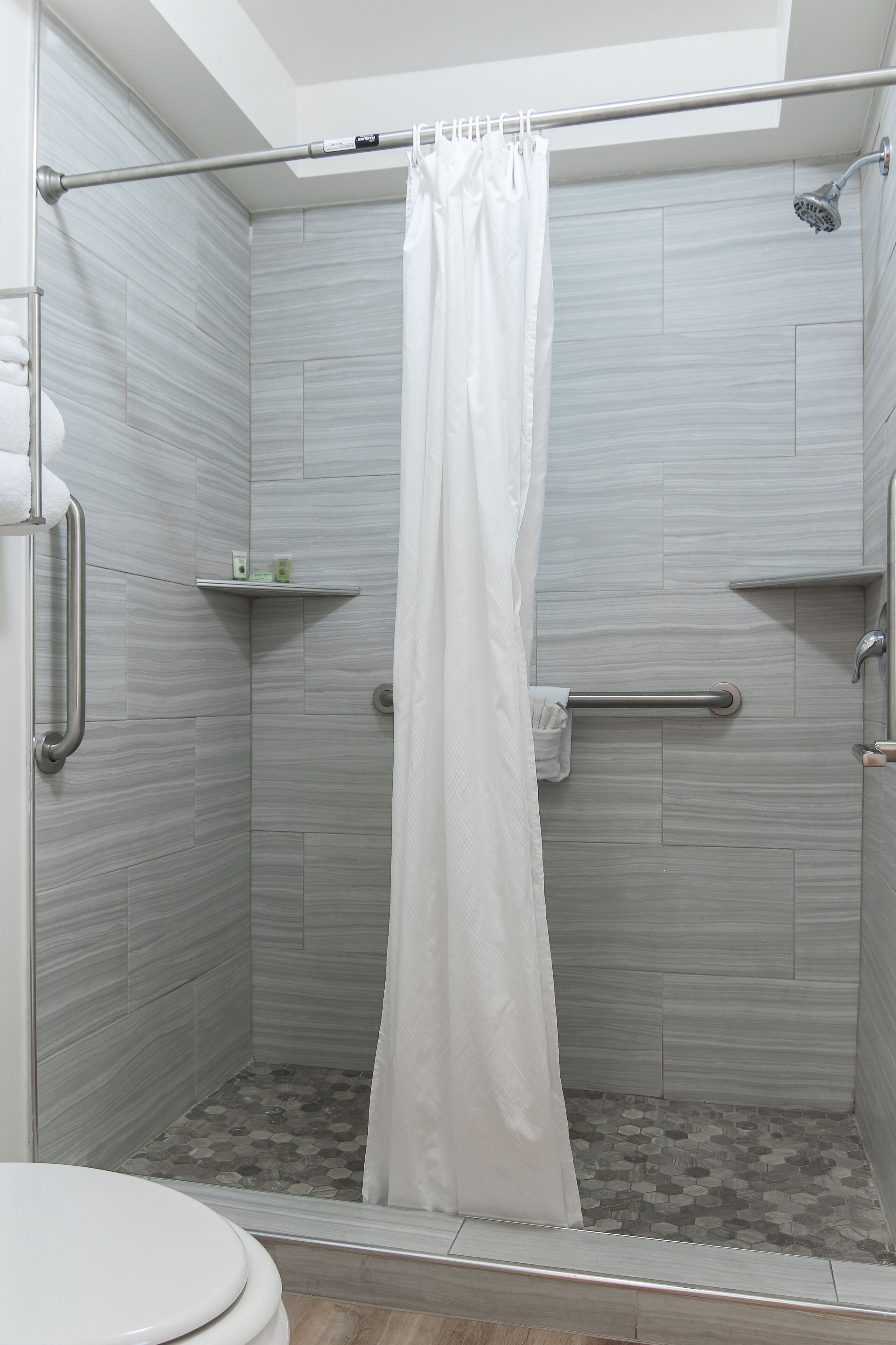 A shower area with a white shower curtain, grab bars, and a small shelf with toiletries.