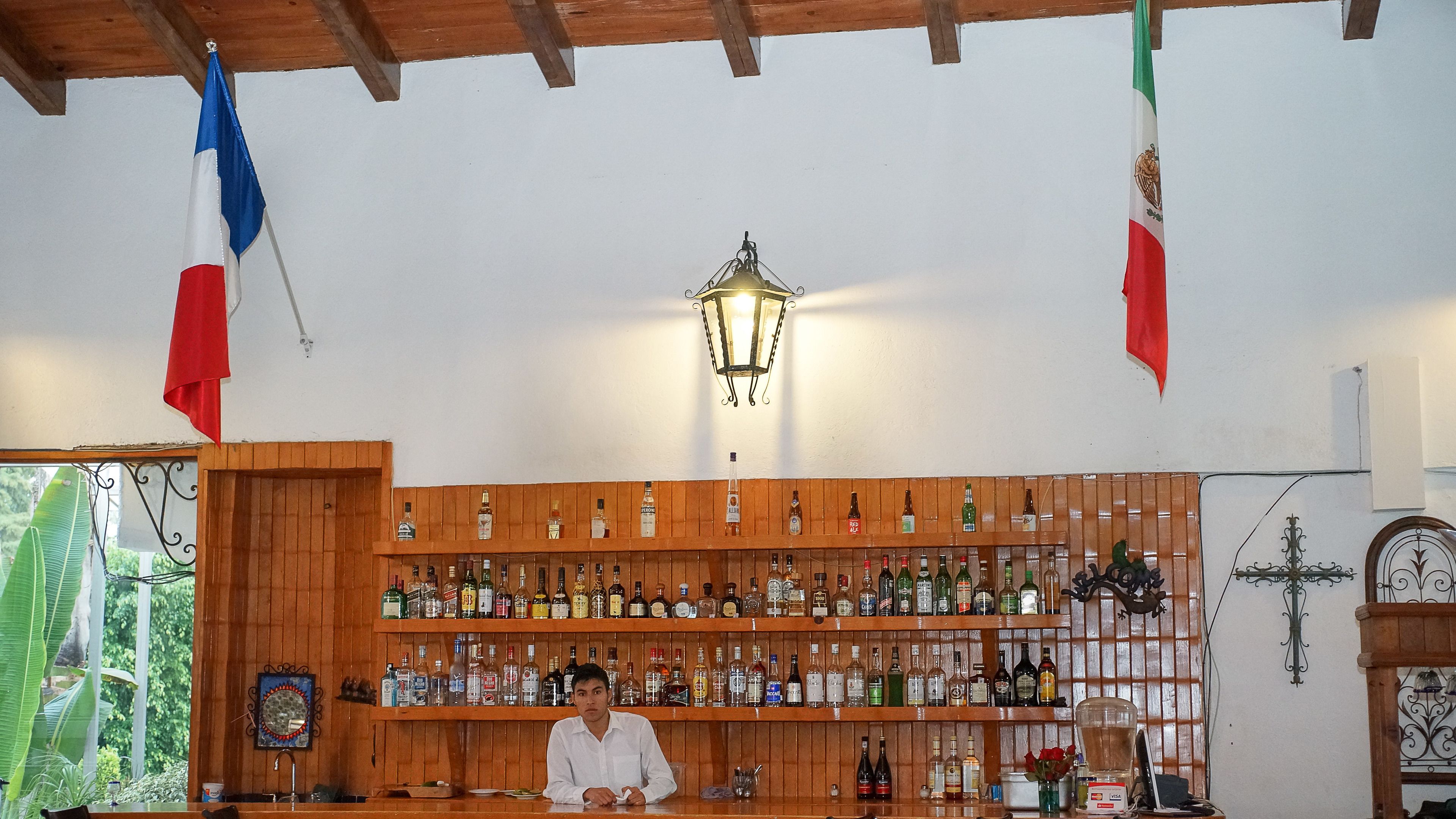 Een bar met houten planken vol verschillende drankflessen, een hanglamp en twee vlaggen.