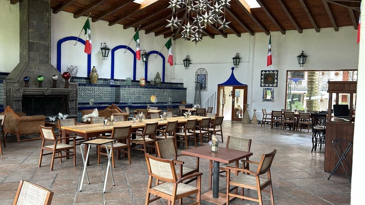 Un amplio espacio exterior con zona de comedor, varias mesas y sillas de madera, una chimenea de piedra y banderas decorativas.