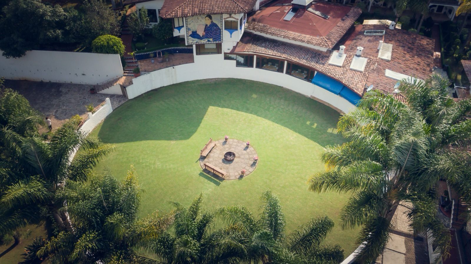An aerial view of a courtyard with a central circular area, surrounded by buildings with tiled roofs and a white wall.
