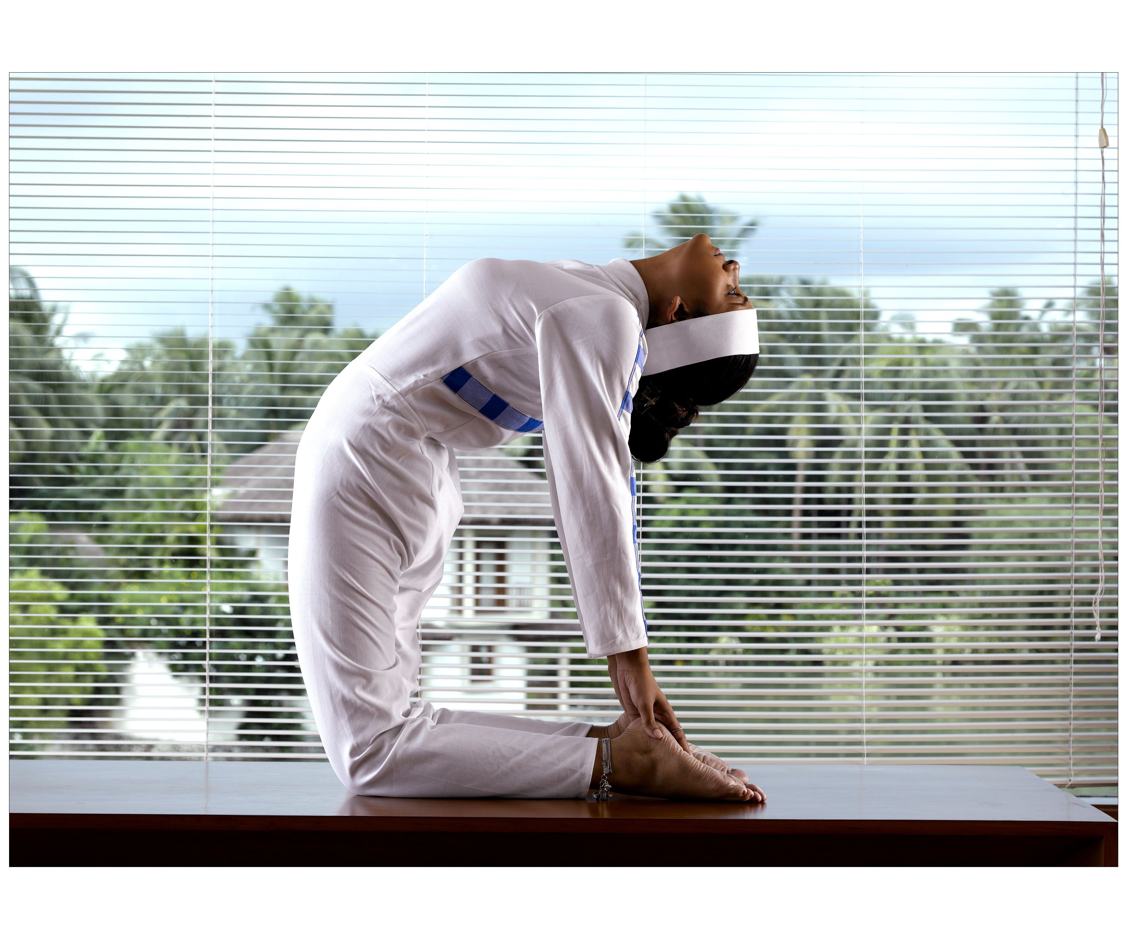 A person in a white outfit performing a yoga pose on a wooden platform by a window with blinds.