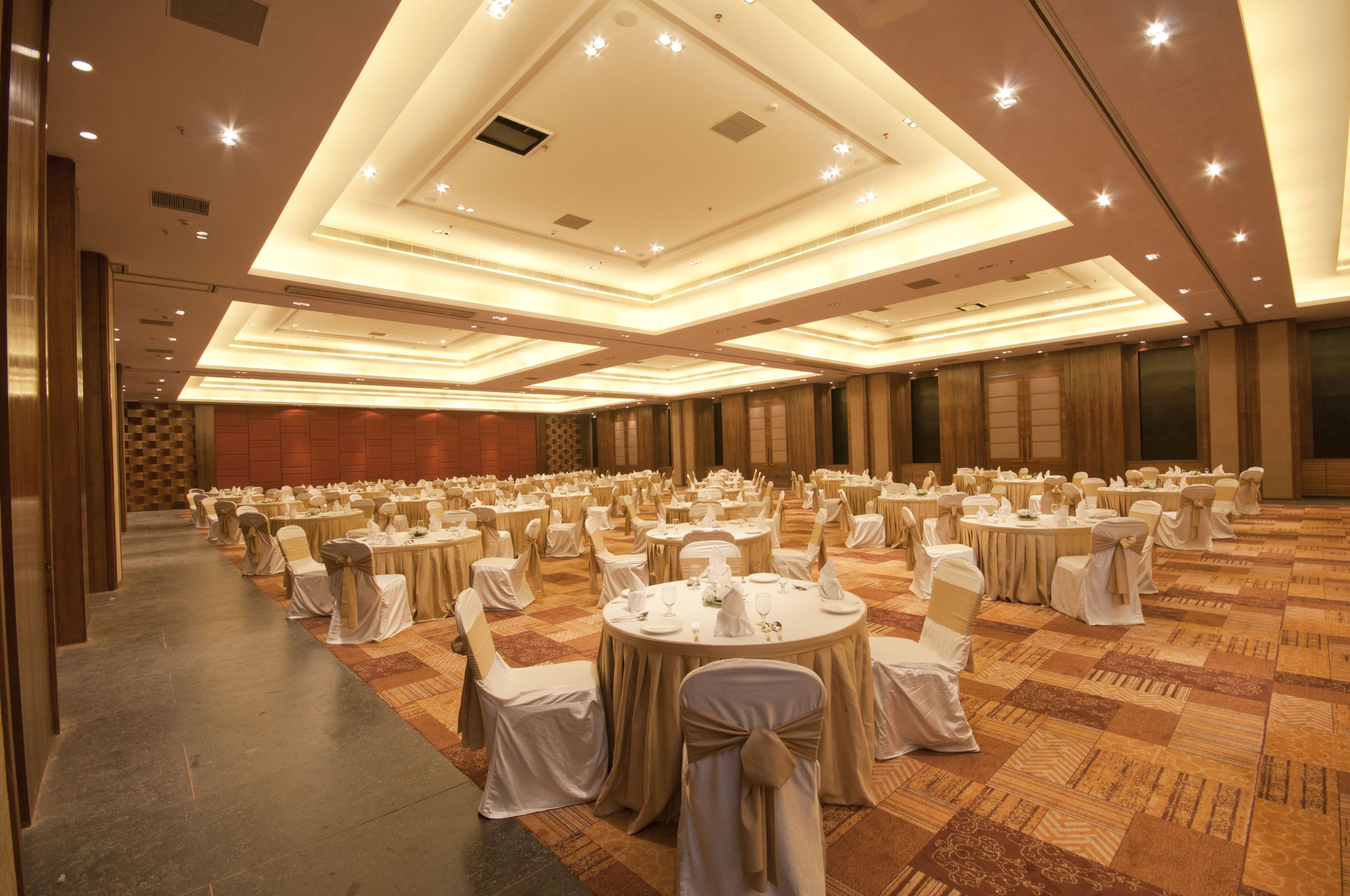 A large banquet hall with round tables set for events, featuring a carpeted floor and a high ceiling with recessed lighting.
