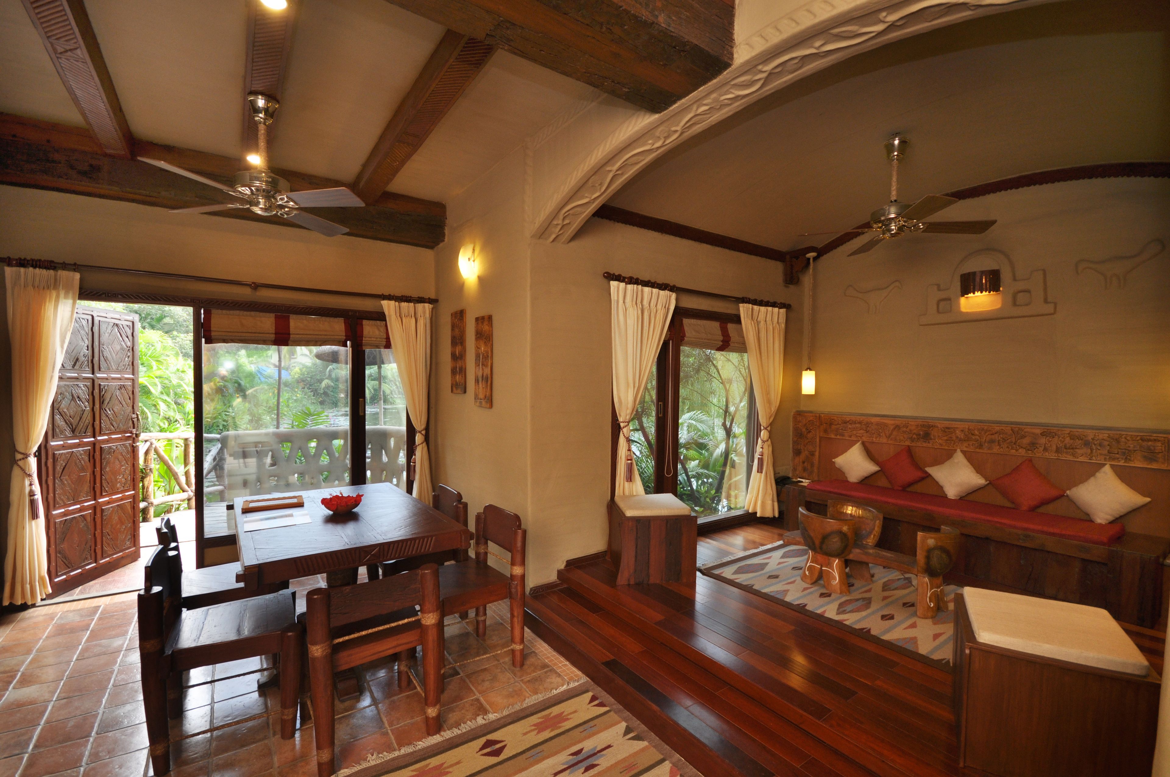 Una habitación con una mesa de comedor de madera, sillas, un banco con cojines, un ventilador de techo y ventanas grandes con cortinas.