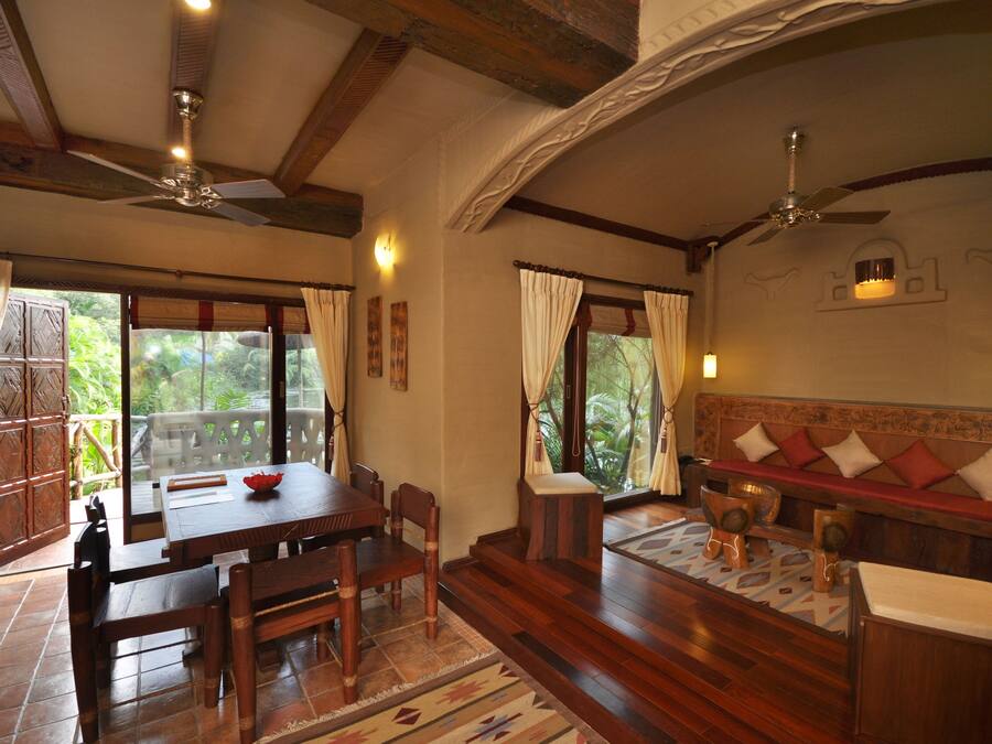 Una habitación con una mesa de comedor de madera, sillas, un banco con cojines, un ventilador de techo y ventanas grandes con cortinas.