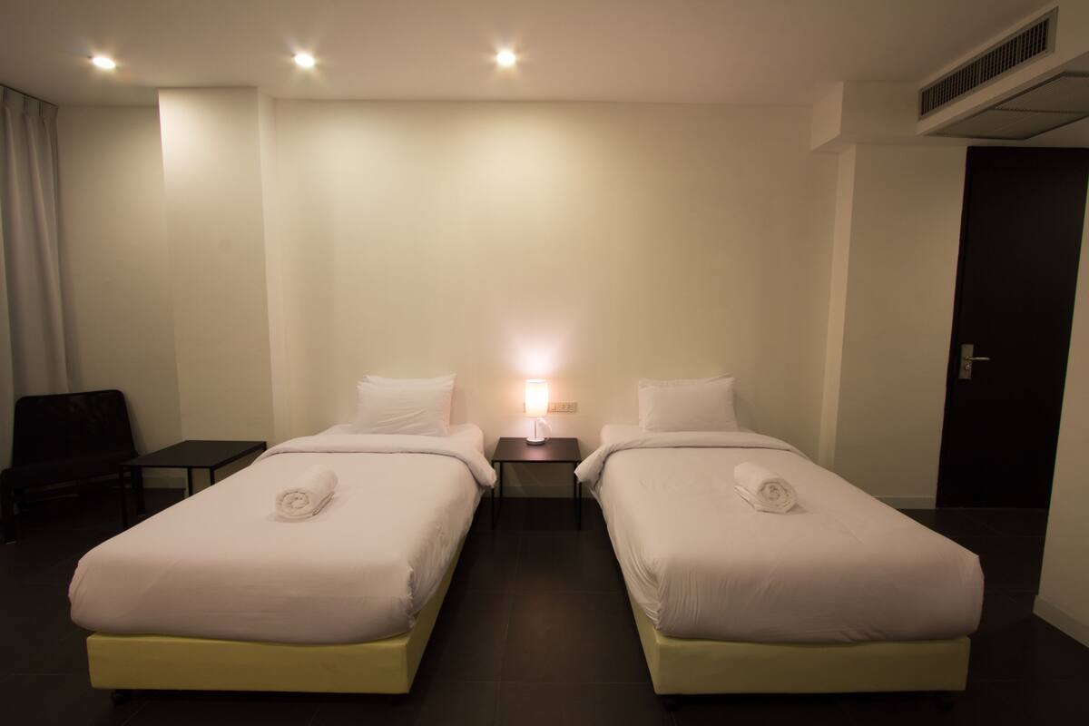 A hotel room with two single beds, a nightstand with a lamp, a chair, and a door.