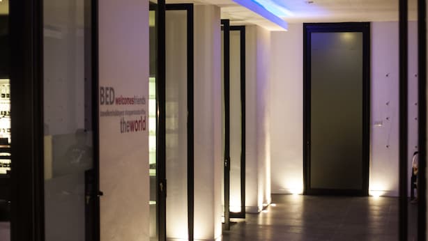 Un couloir d’hôtel moderne avec des portes en verre, une enseigne indiquant « BED accueille ses amis du monde entier » et un plafond éclairé de lumière bleue.