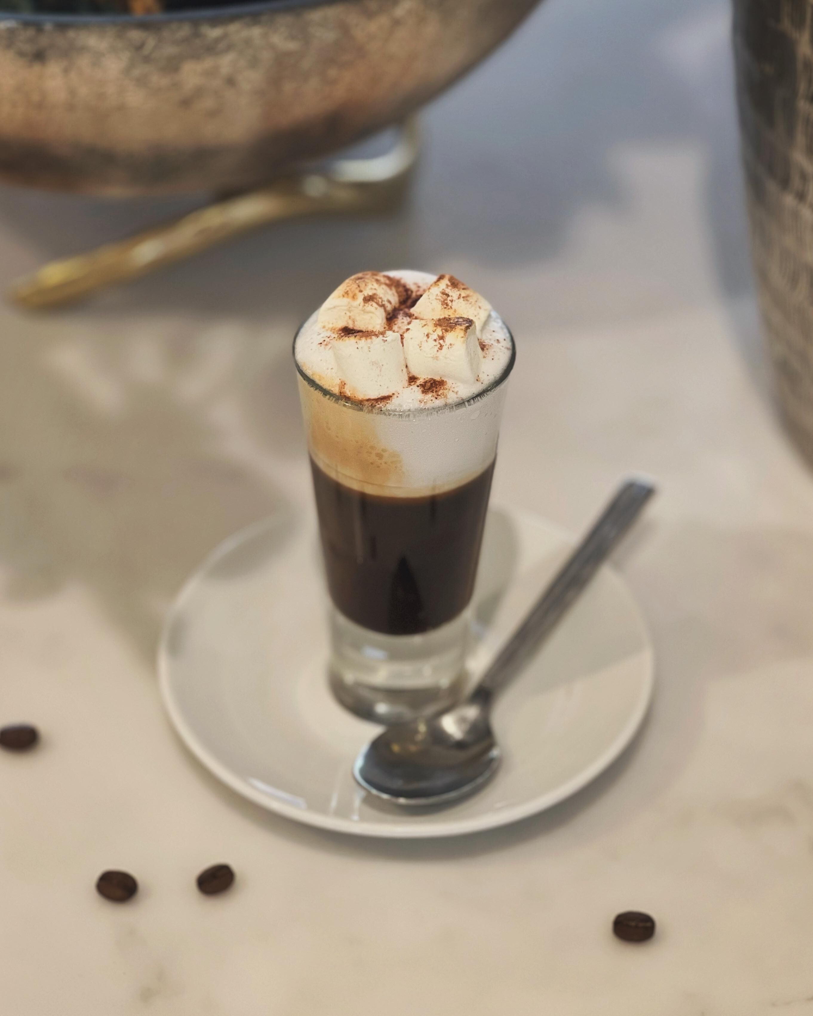 A tall glass with a layered beverage topped with whipped cream and cocoa powder, placed on a white plate with a spoon beside it.