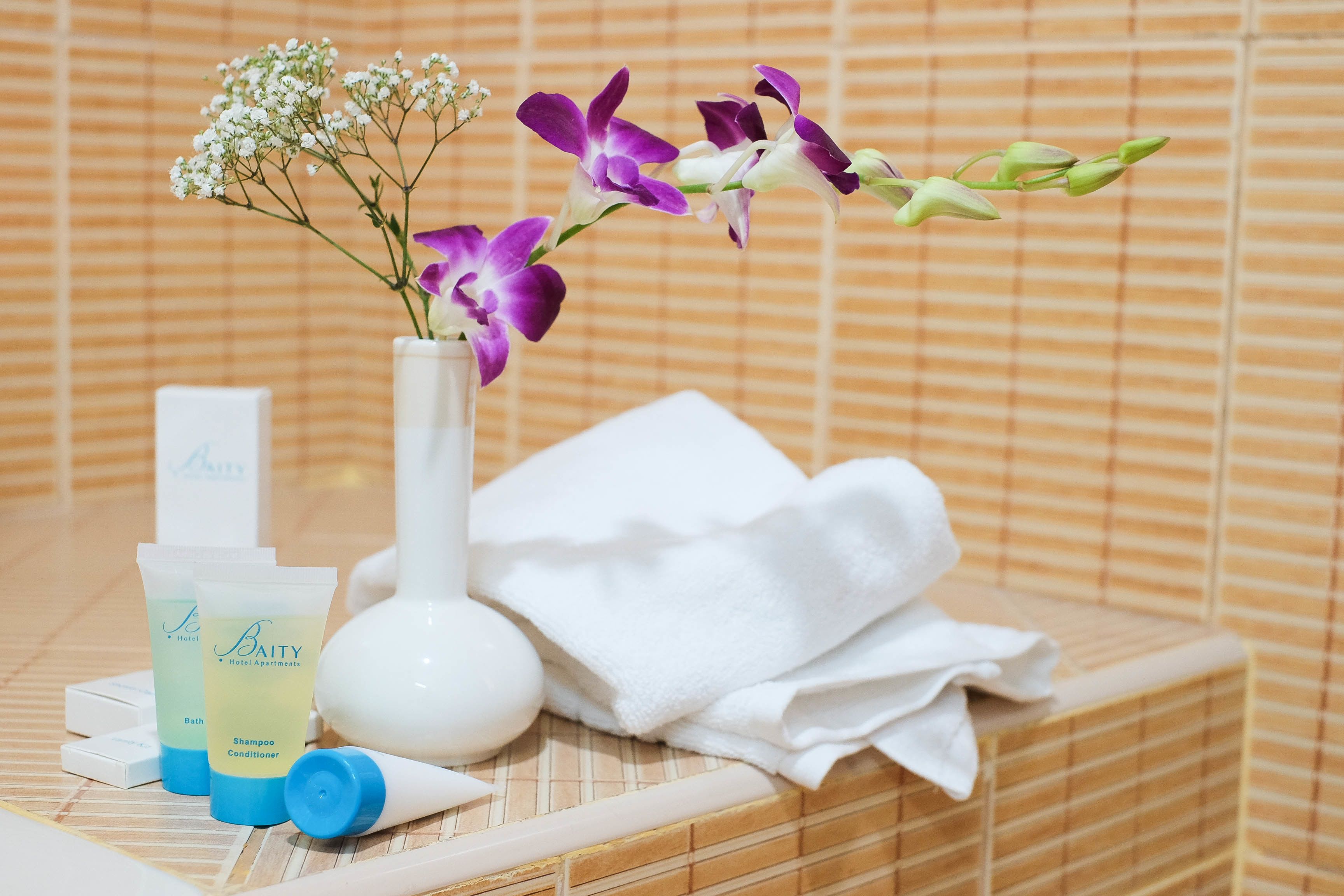 A bathroom with a white vase containing purple and white flowers, a white towel, and two bottles of Baity Hotel Apartments shampoo and conditioner.