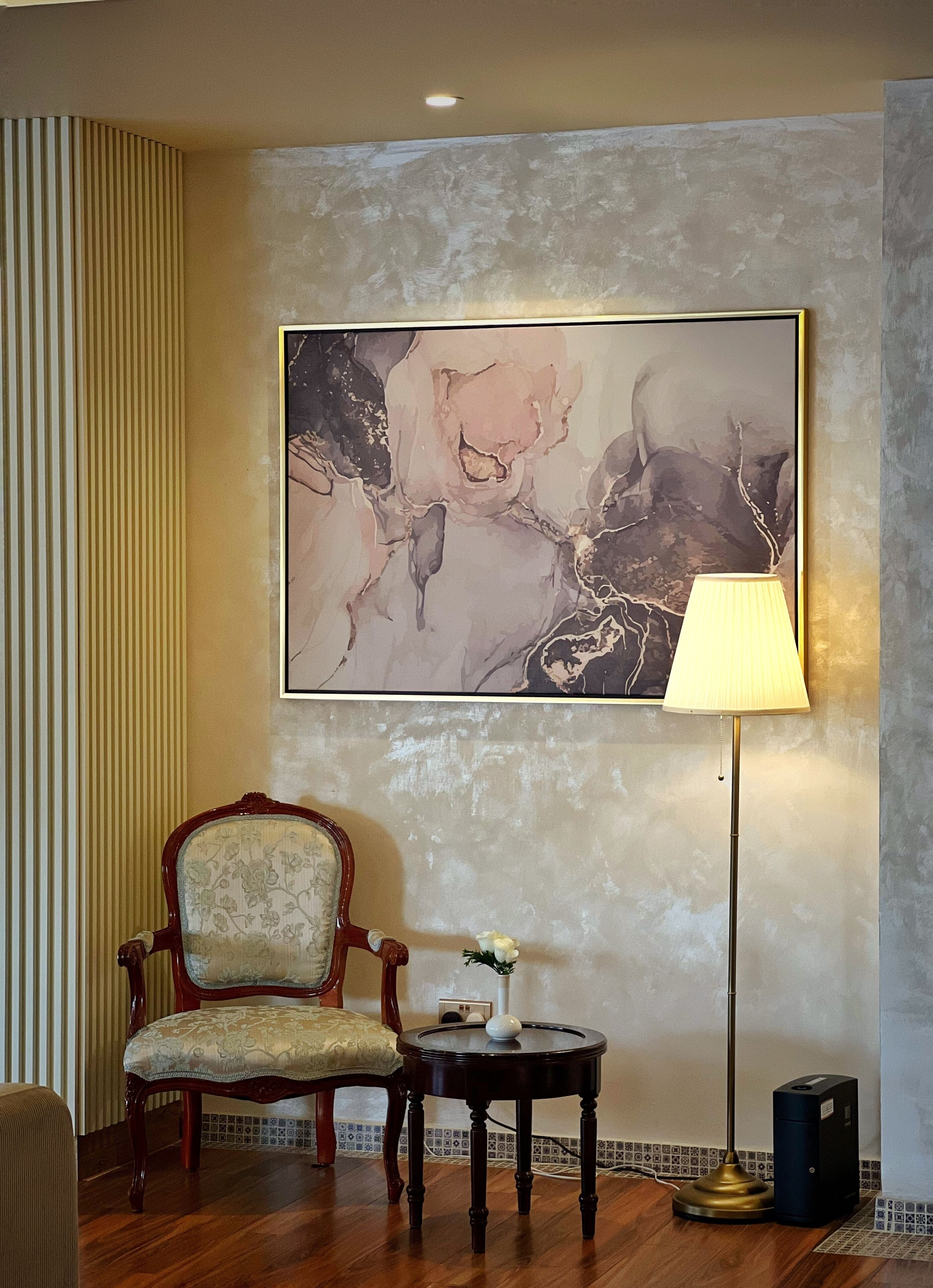 Een kamer met een groot abstract schilderij aan de muur, een houten stoel, een klein tafeltje met een vaas, een vloerlamp en een speaker.