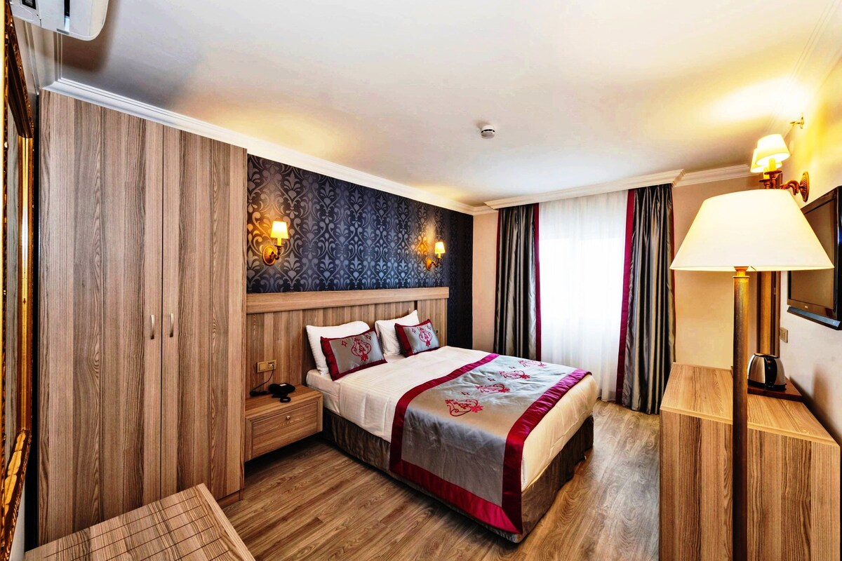 A hotel room with a wooden wardrobe, a bed with a red and white bedspread, two bedside lamps, and a nightstand.